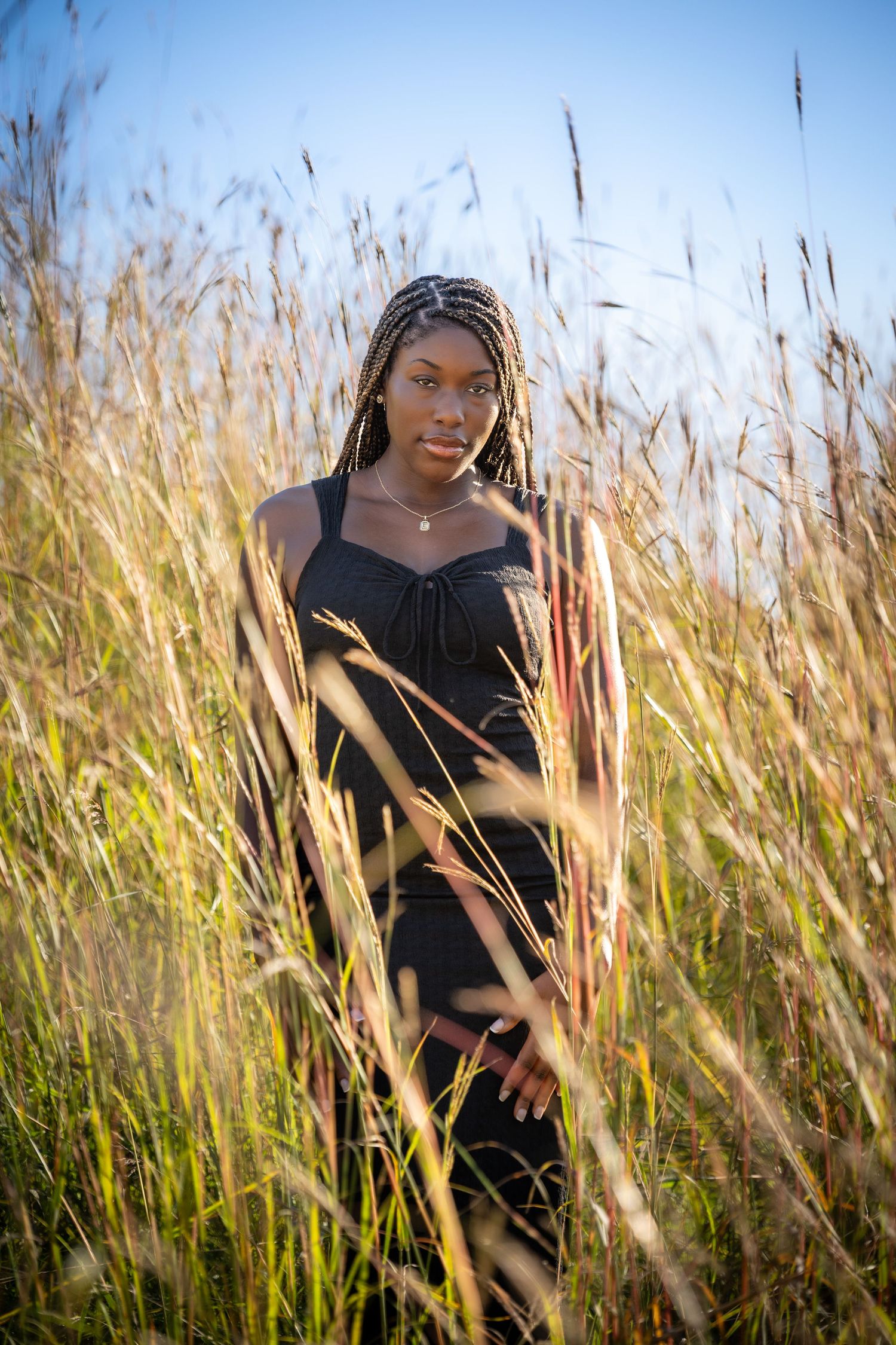 Young Black woman with braids wearing a black dress standing in tall golden grass under a blue sky.