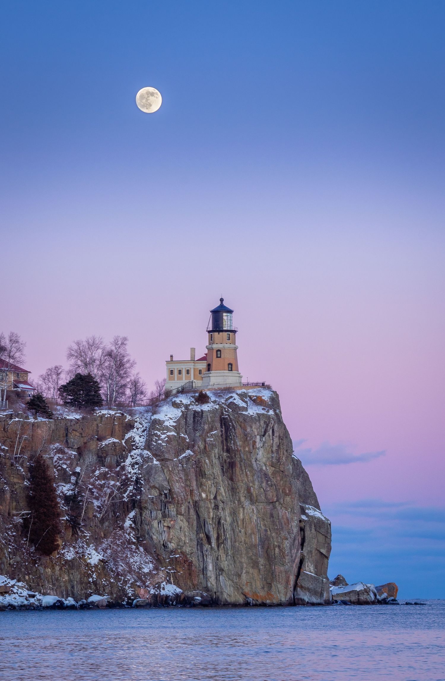 Split Rock Lighthouse perched on snowy cliffs above Lake Superior under a full moon at twilight.