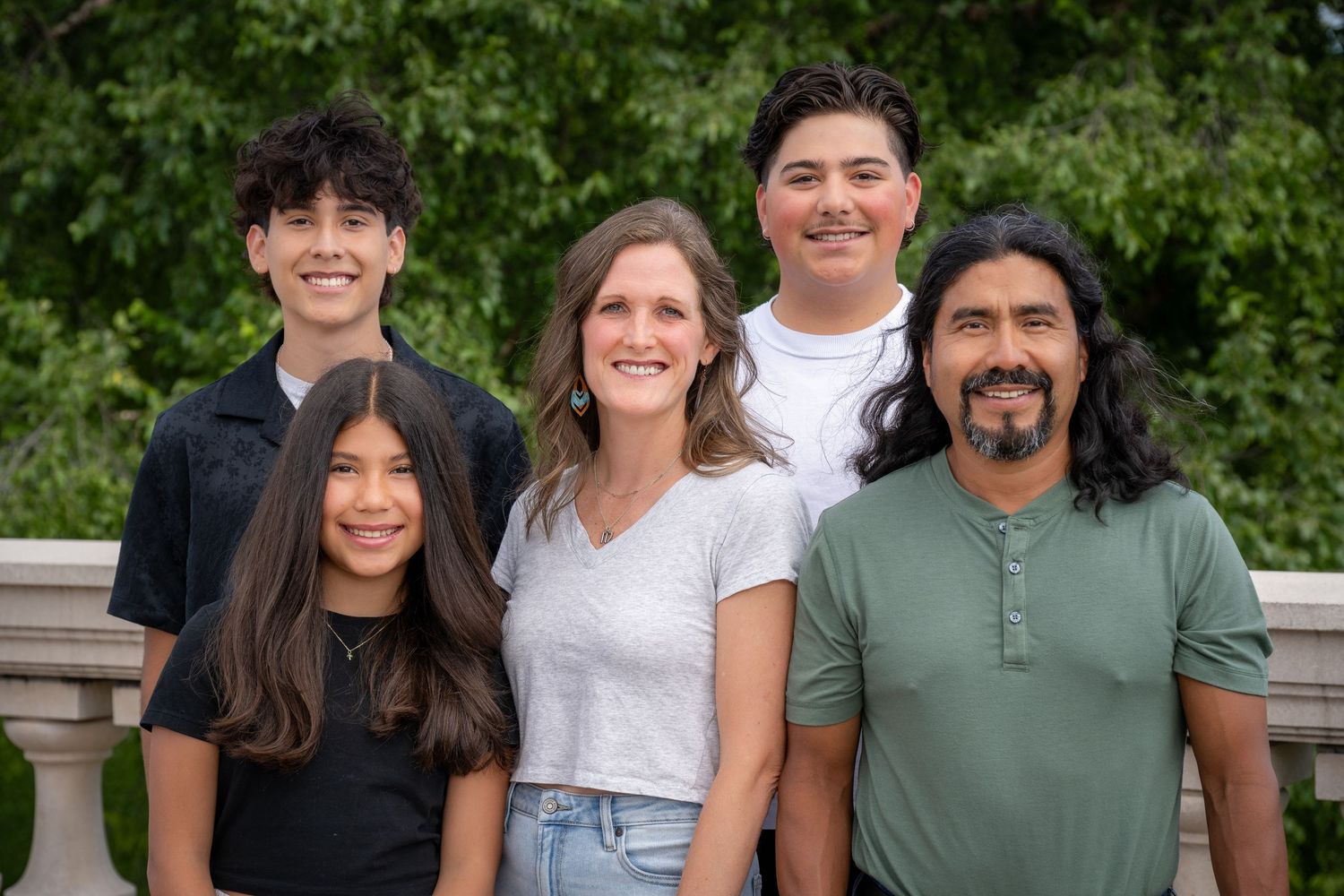 Happy family of five smiling together outdoors with lush green trees in the background.