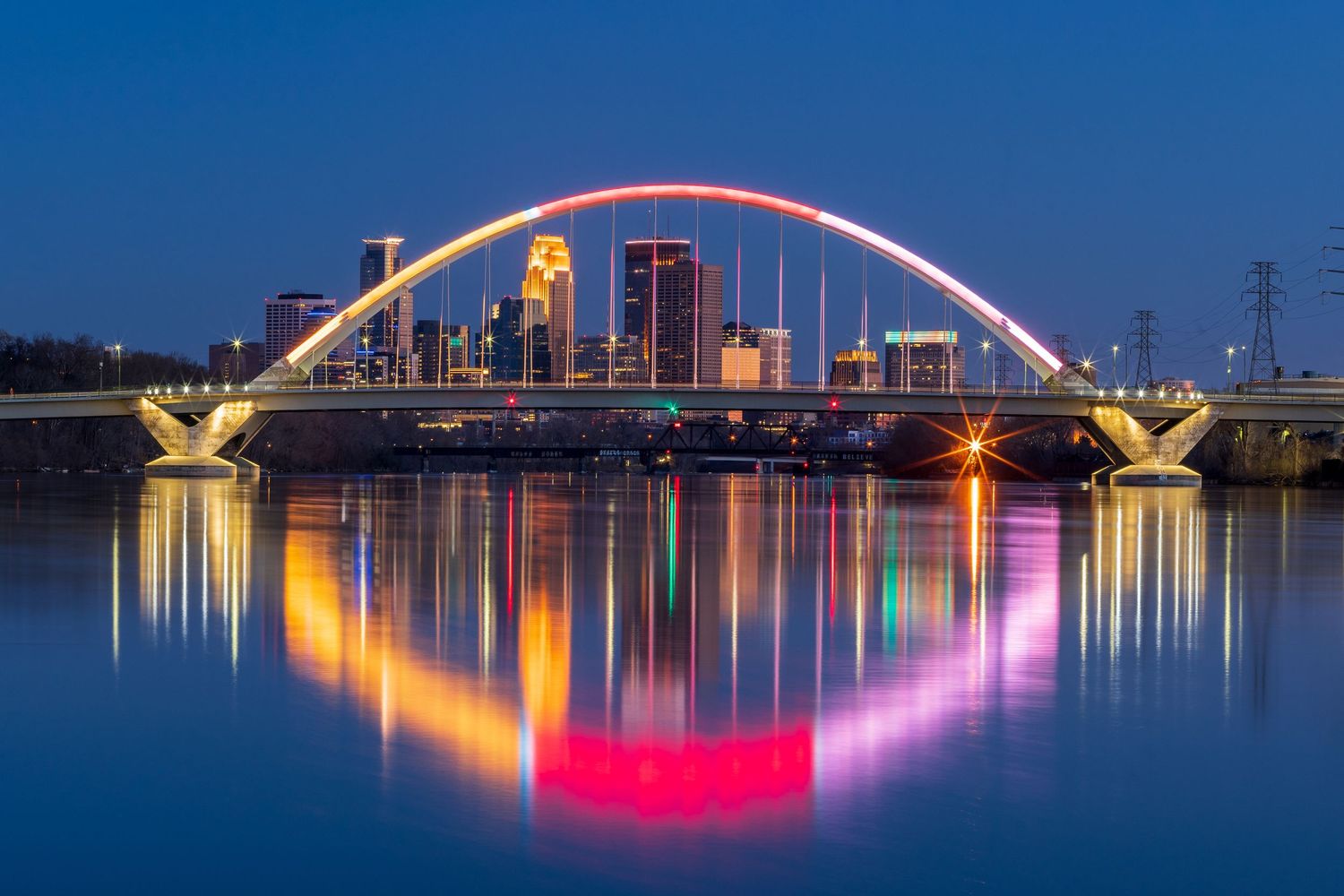 Minneapolis skyline reflected in river at blue hour with illuminated arch bridge.