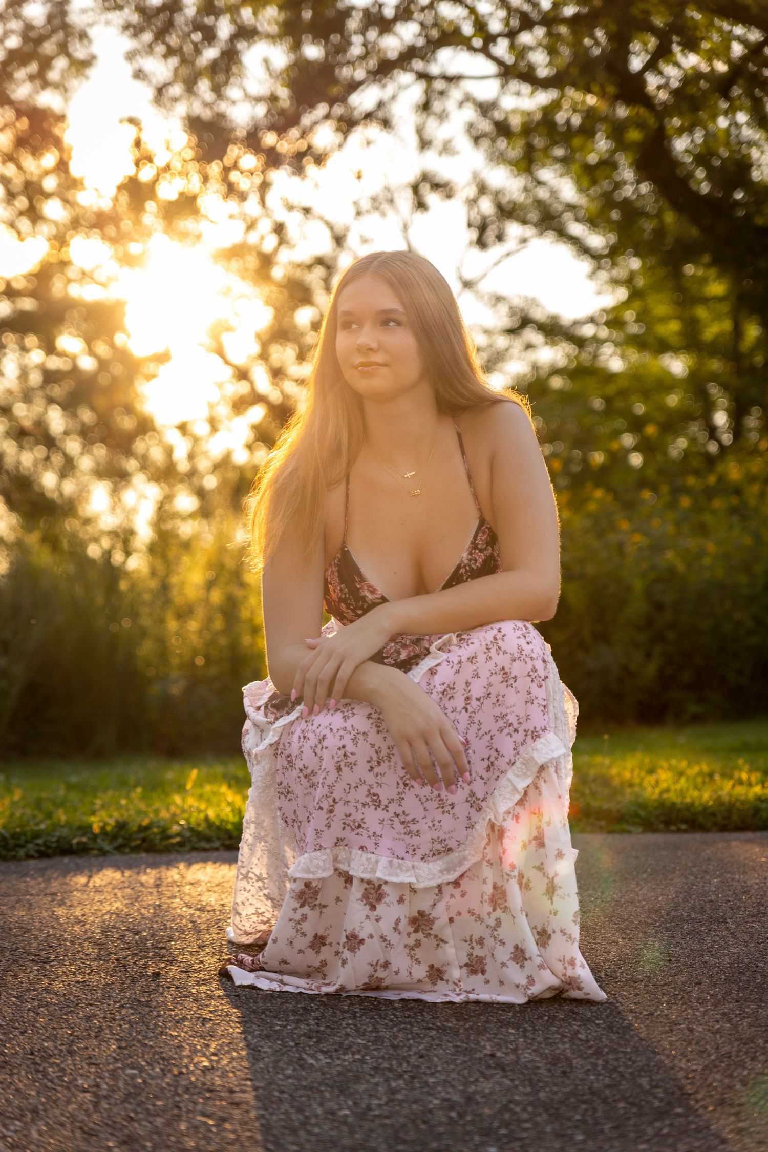 Young woman in floral dress sitting on pavement at golden hour sunset with trees in background.