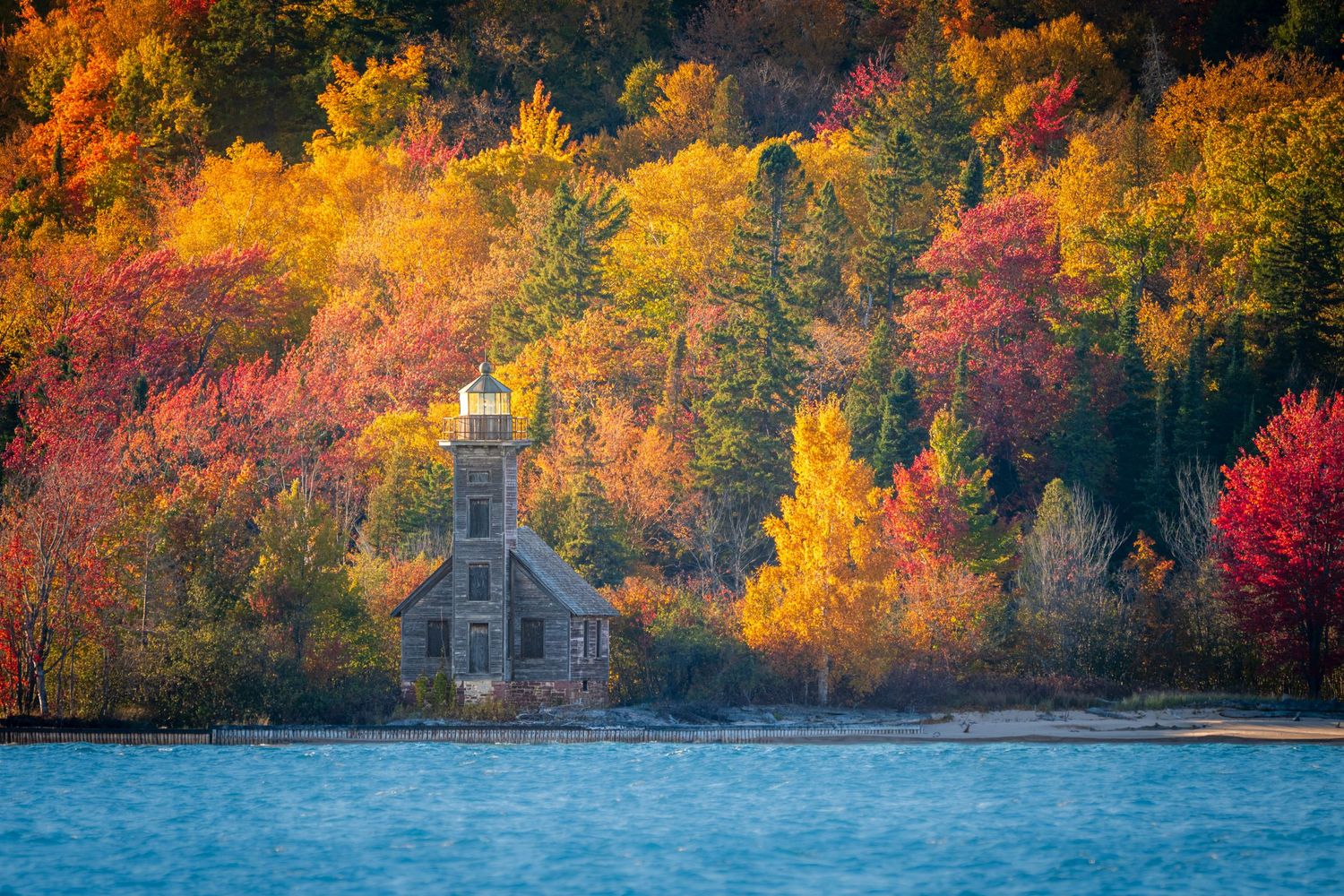 Historic lighthouse surrounded by vibrant autumn foliage in red, orange, and yellow hues along a calm lakeshore.