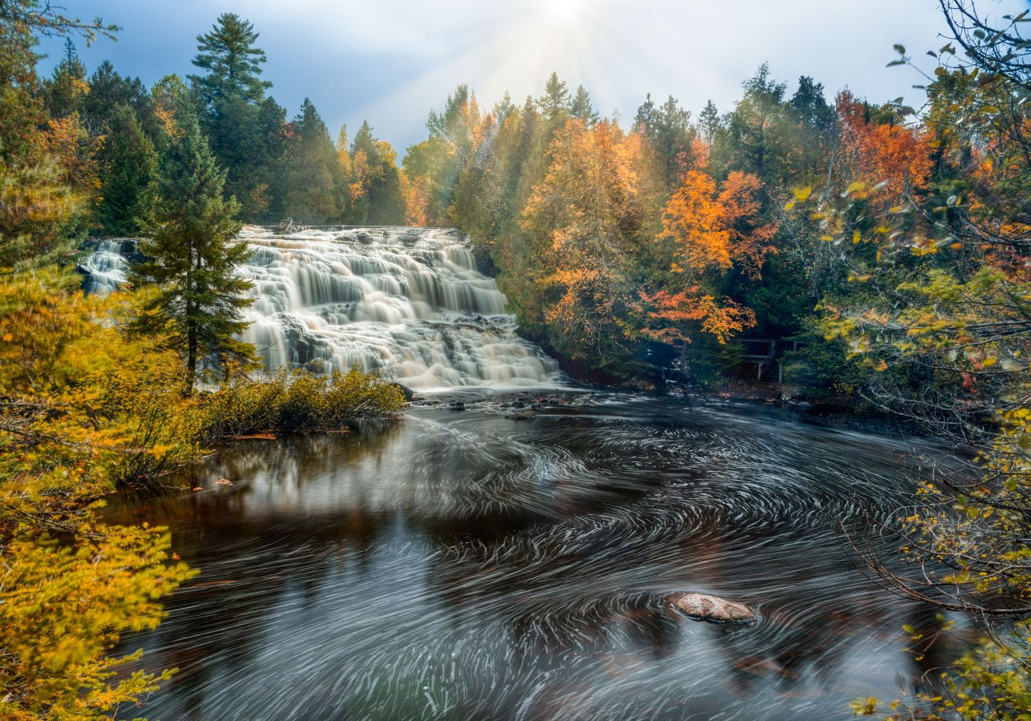 Stunning waterfall cascading into a swirling river pool surrounded by colorful autumn trees with sunbeams breaking through.