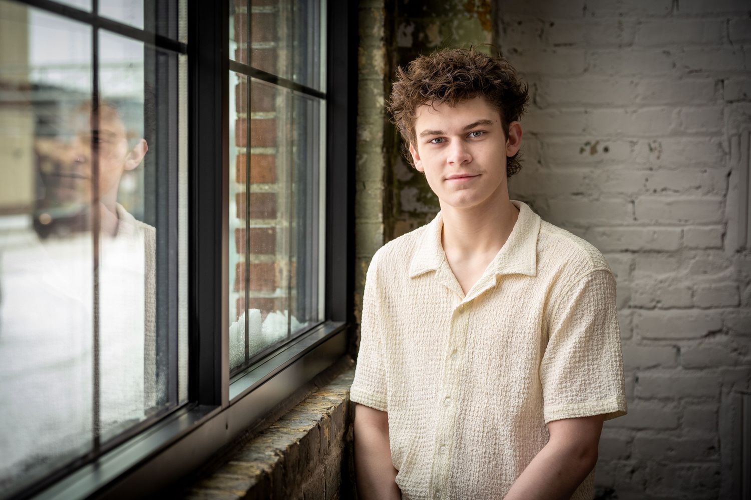 Young man in a beige shirt sitting by a large window in a rustic brick-walled interior.