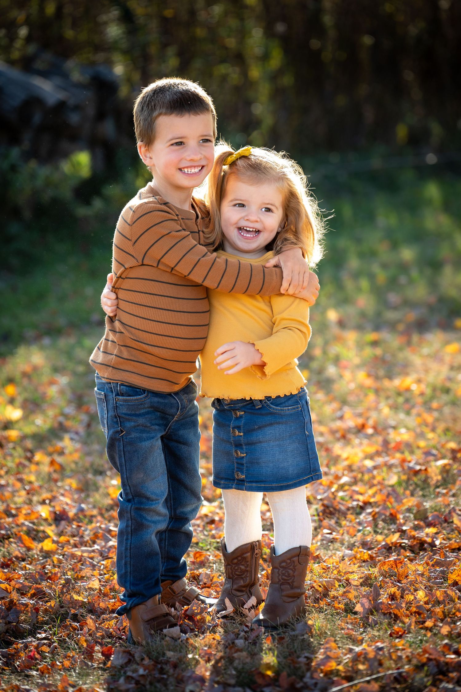 Two happy young siblings hugging outdoors in a sunlit autumn park covered in fallen orange and red leaves.