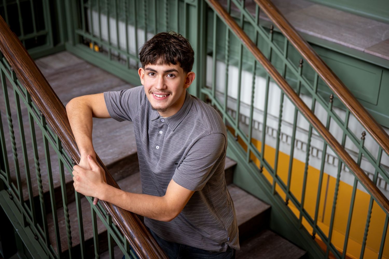 Smiling young man leaning on a railing on a staircase with green and yellow painted walls indoors.