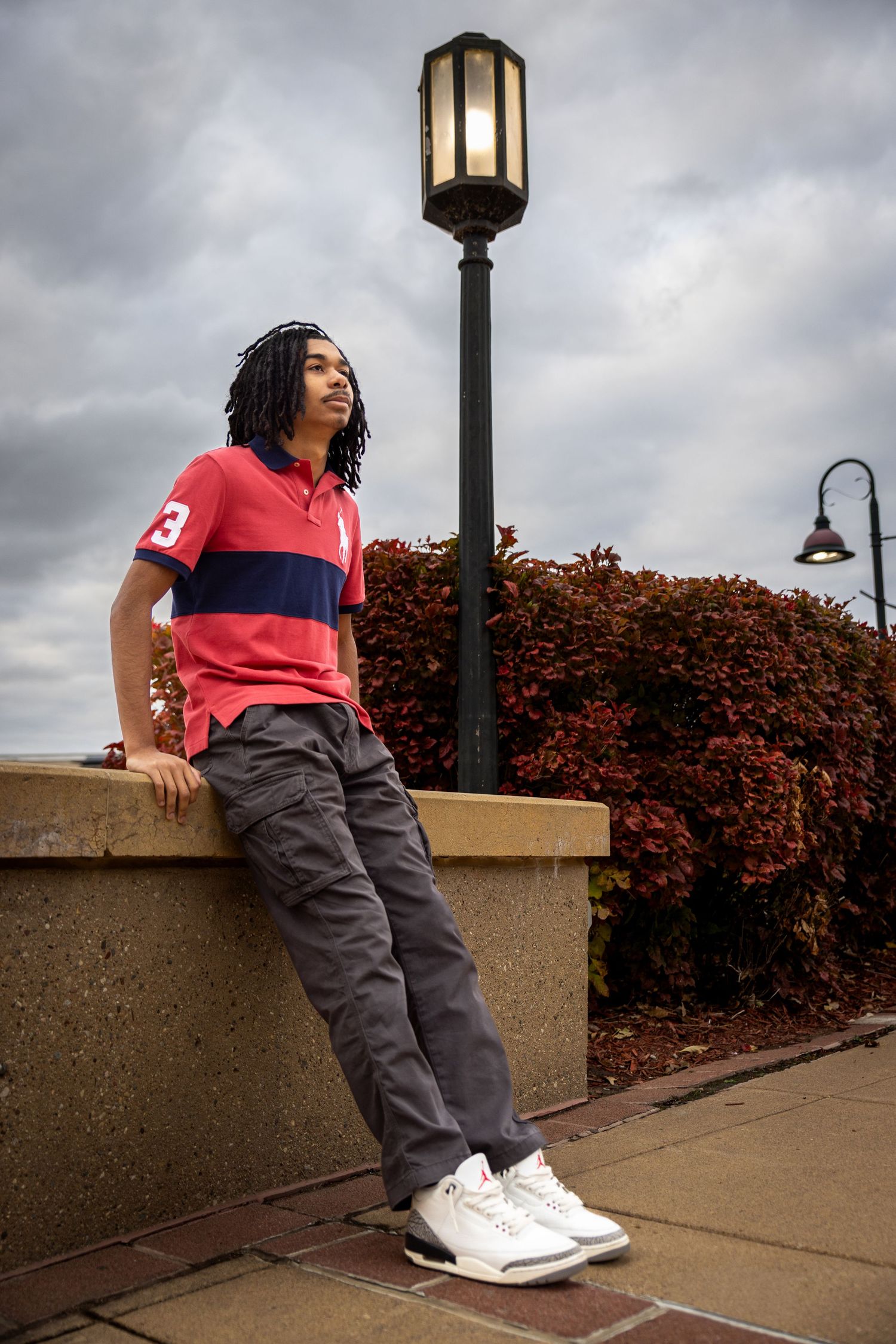 Young man in red polo and gray cargo pants leaning against a wall near a lamp post on a cloudy day.
