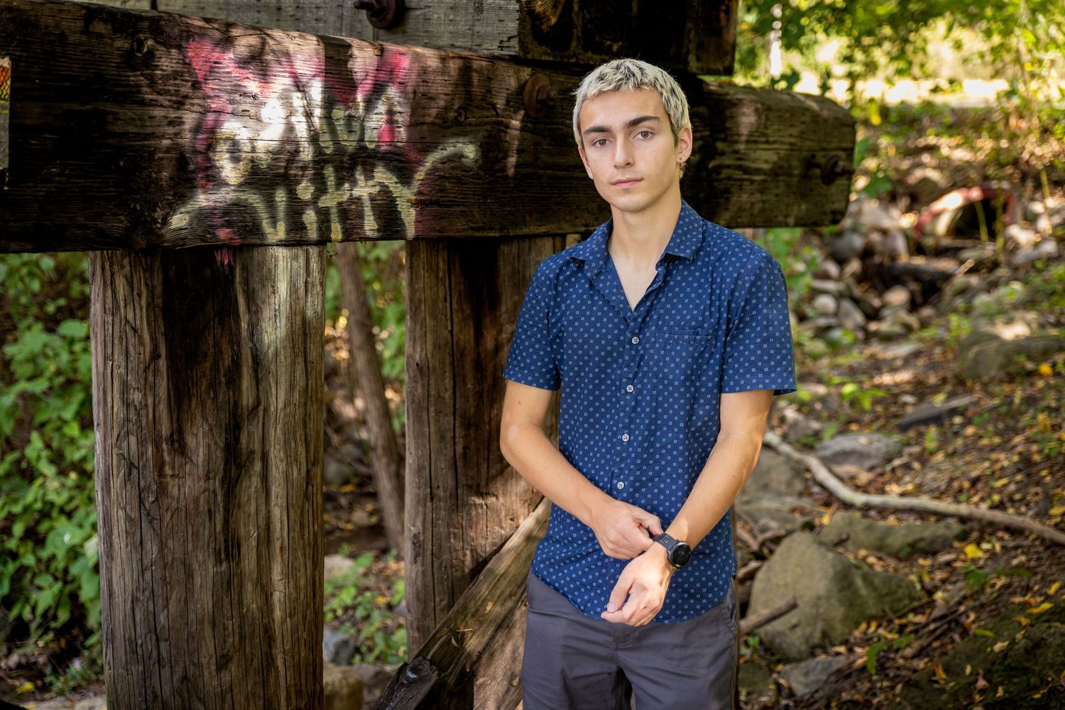 Young man in a navy blue shirt standing near graffiti-covered wooden beams in a wooded area.