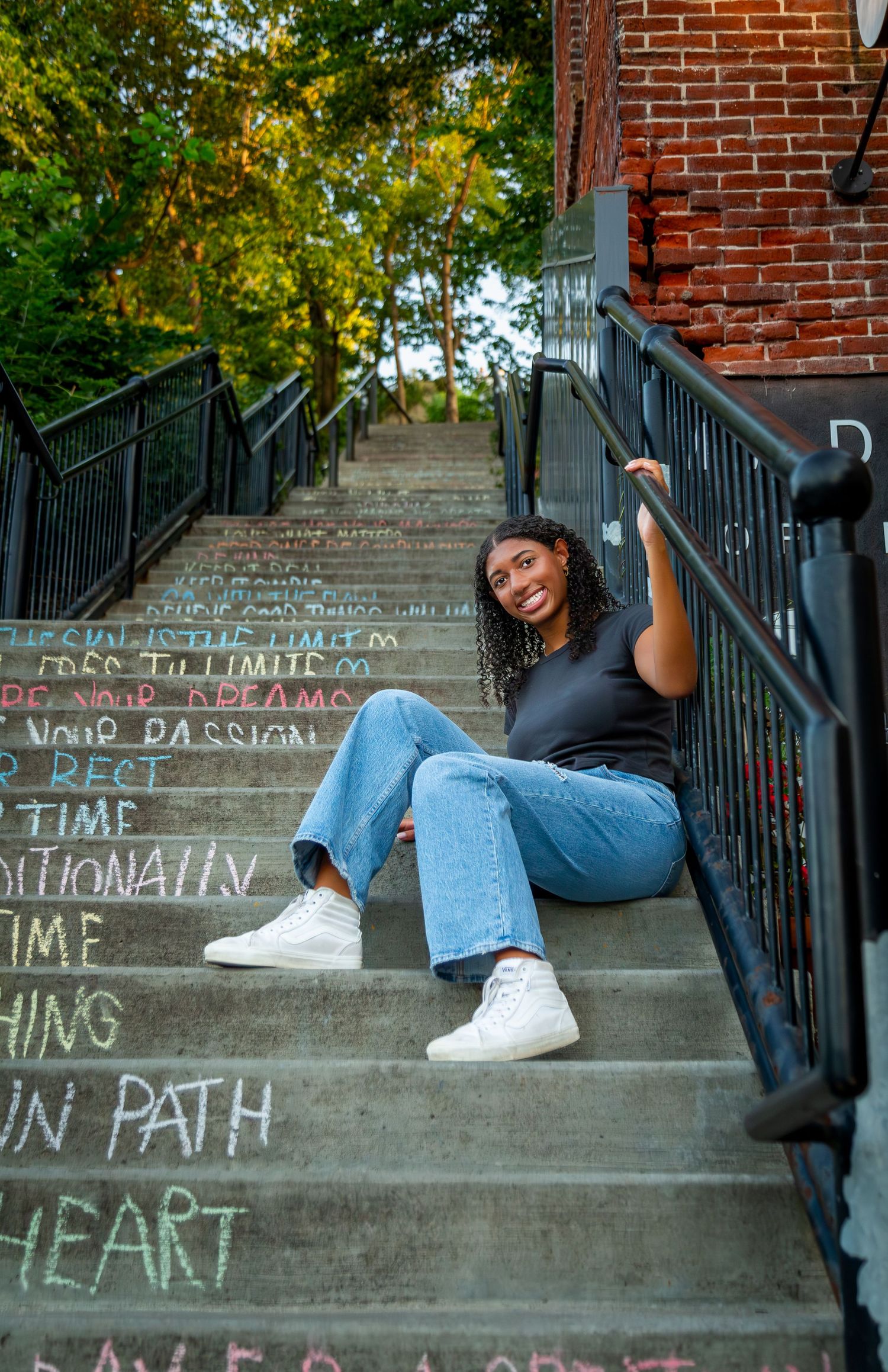 Young woman smiling on chalk-written steps near brick building, wearing black top, jeans, and white sneakers.