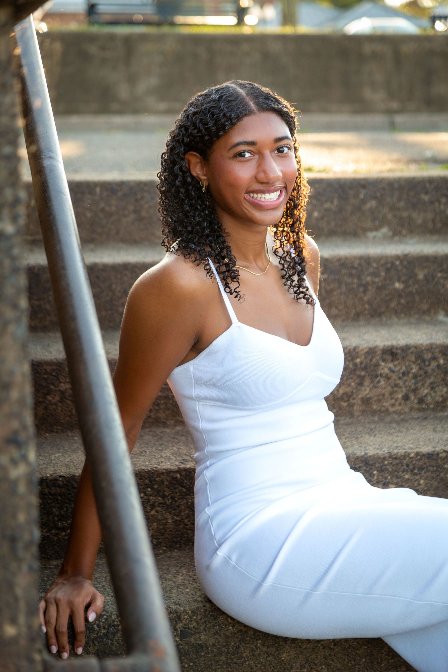 Smiling young woman in white outfit sits on stone steps in warm sunlight.