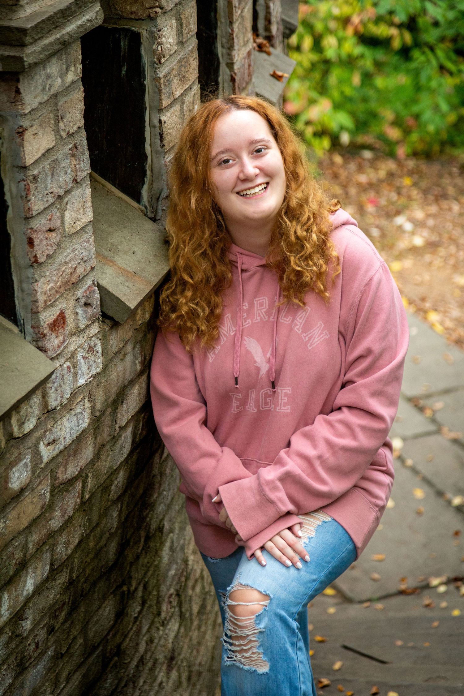 Smiling young woman with curly red hair wearing a pink hoodie and ripped jeans leaning against a brick wall outdoors.