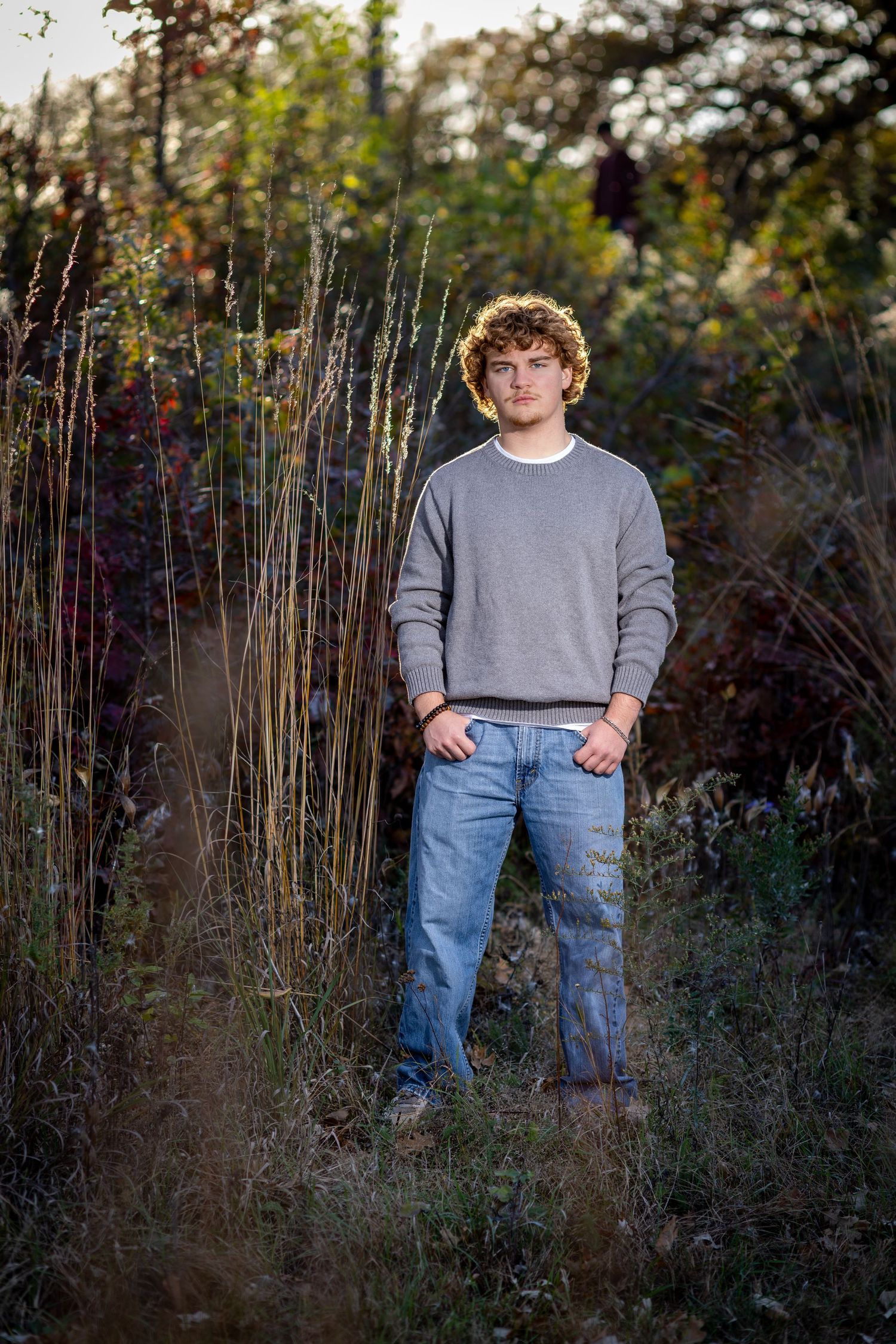 Young man with curly hair in gray sweater and jeans standing outdoors among tall grasses and autumn foliage.