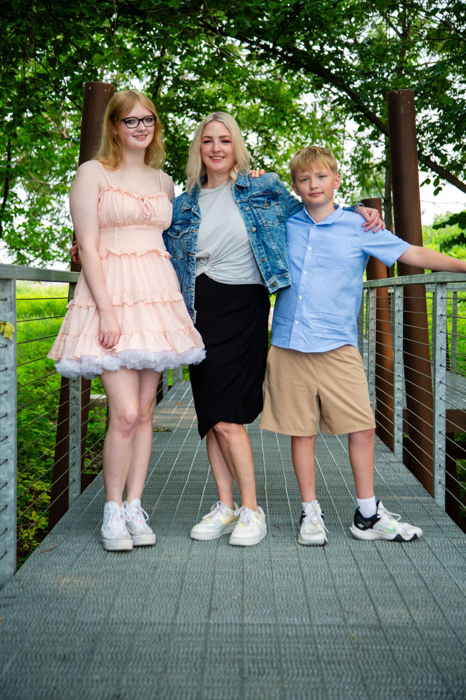 A woman with two children poses on a bridge surrounded by lush green trees on a sunny day.