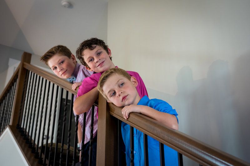Three children lean over a wooden stair railing, smiling down at the camera in a bright modern home.