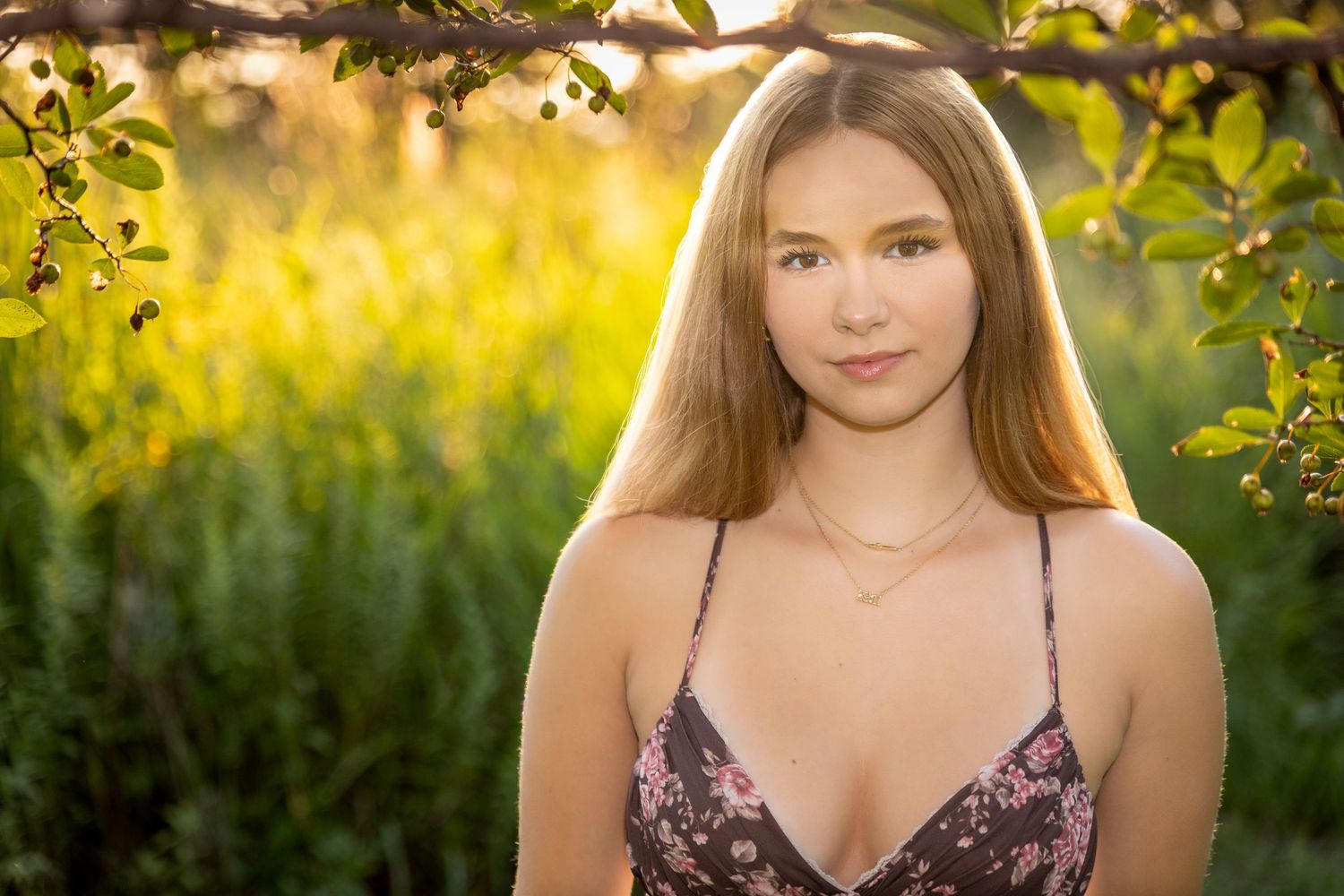 Young woman in floral dress poses outdoors among green foliage in golden hour sunlight.