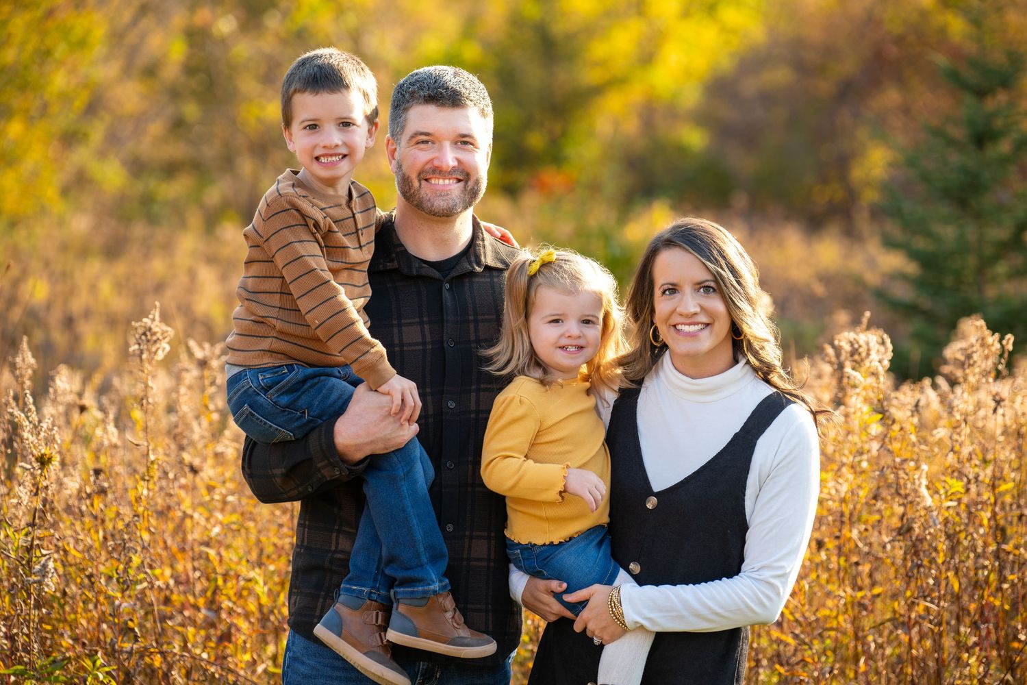 Family of four smiling outdoors in autumn, parents holding young children among golden fall foliage.