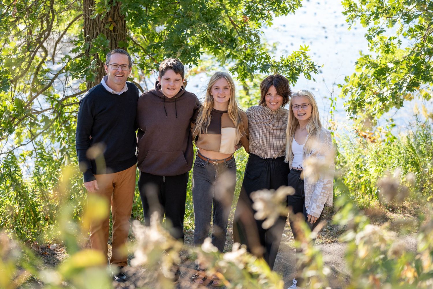 Family of five smiling together outdoors among autumn foliage near a lake on a sunny day.