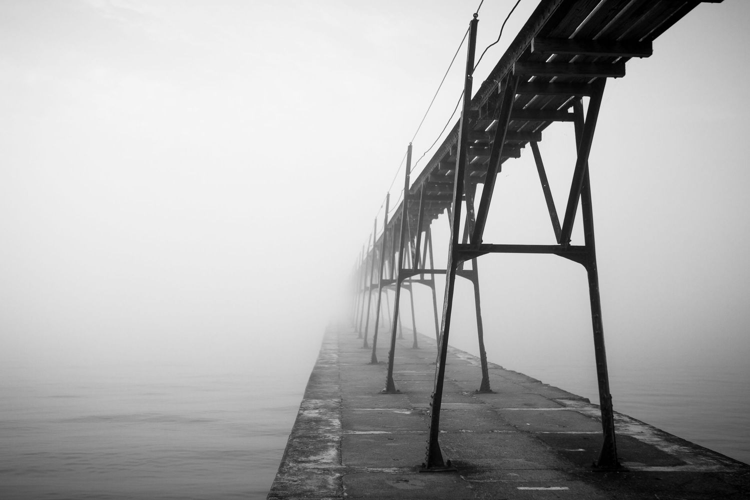 Black and white photo of a foggy pier extending into misty waters with steel support beams.