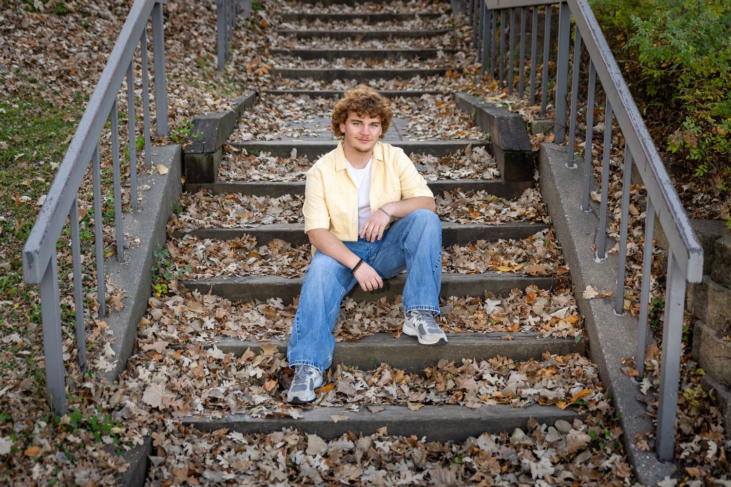 Young man in yellow shirt and jeans sits on outdoor stairs surrounded by fallen leaves.