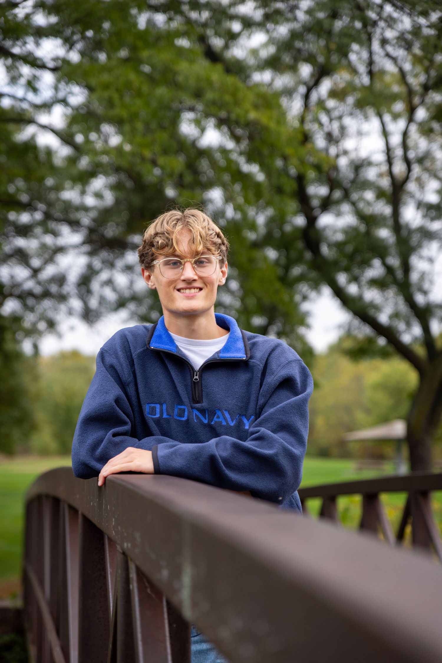 Smiling teen in Old Navy fleece leans on bridge railing in lush green park setting.