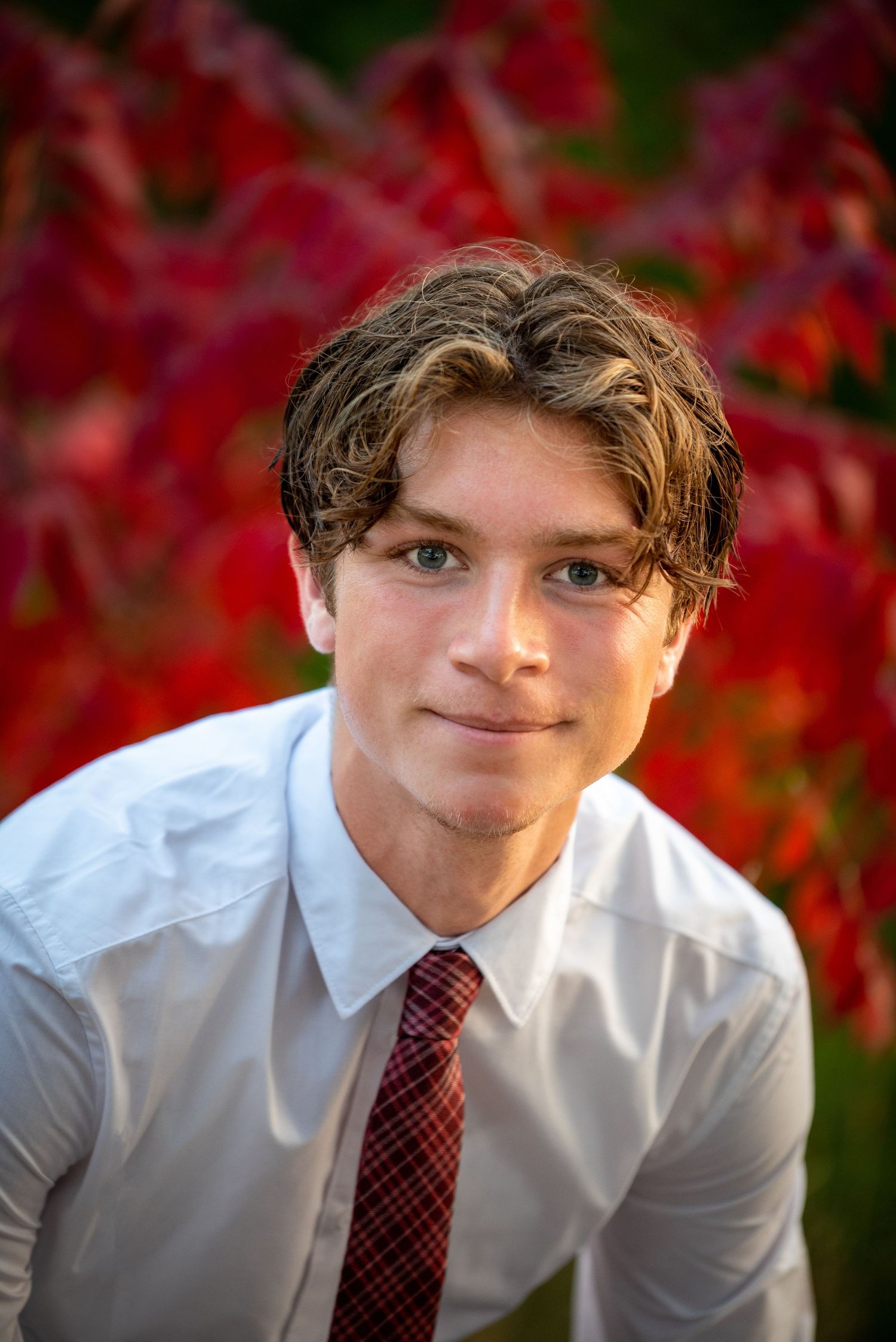 Smiling teenage boy in a white shirt and tie posing in front of vivid red autumn foliage.