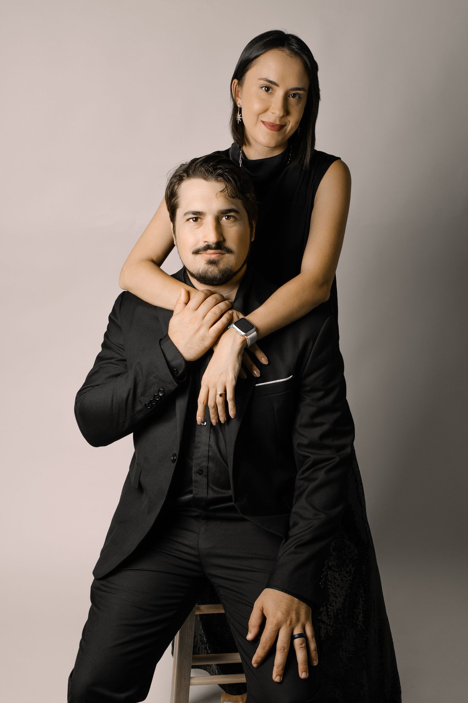 A couple in matching black outfits poses together, woman embracing man from behind in a studio setting.