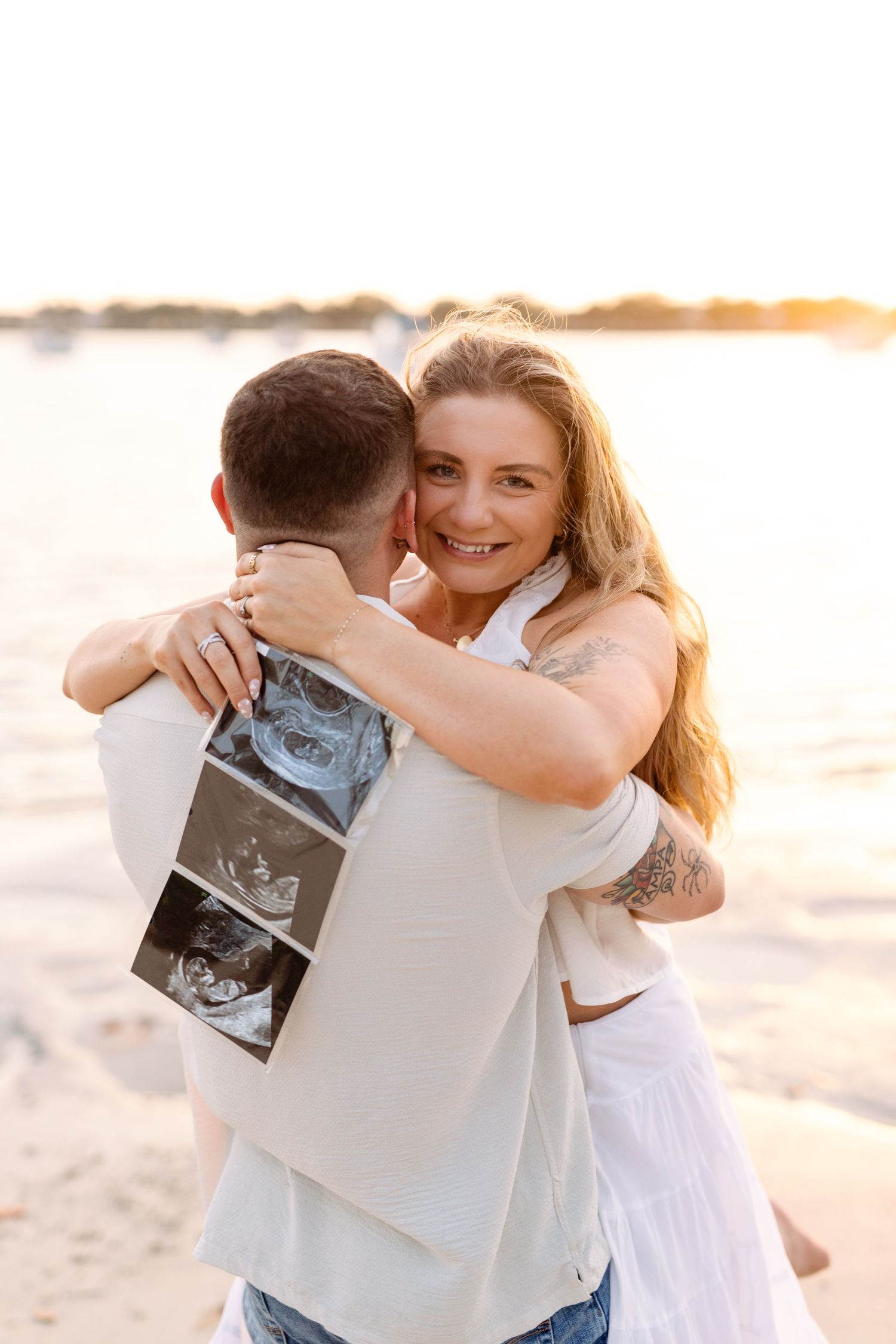 Smiling woman hugs man on beach at sunset while holding ultrasound photos, announcing pregnancy at Davis Island.