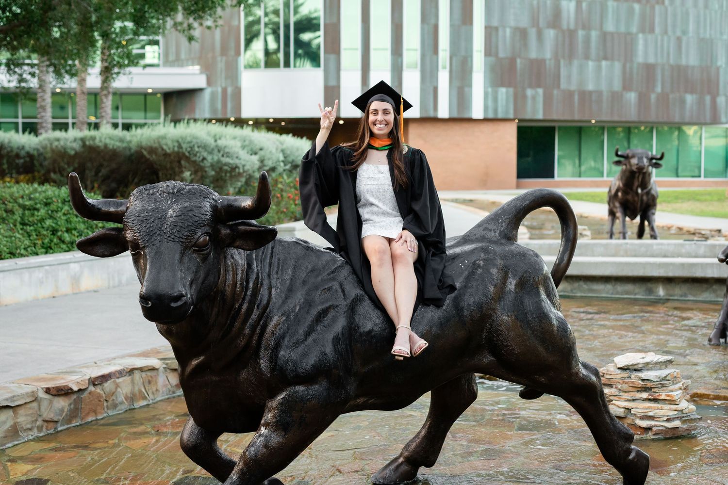 A smiling graduate in cap and gown sits atop a bronze bull statue on a university campus at the University of South Florida.