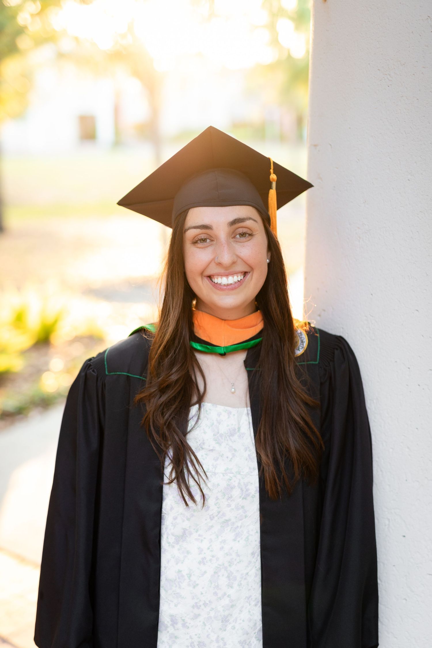 Smiling female graduate in black cap and gown leaning against a white column in warm sunlight at the University of South Florida.
