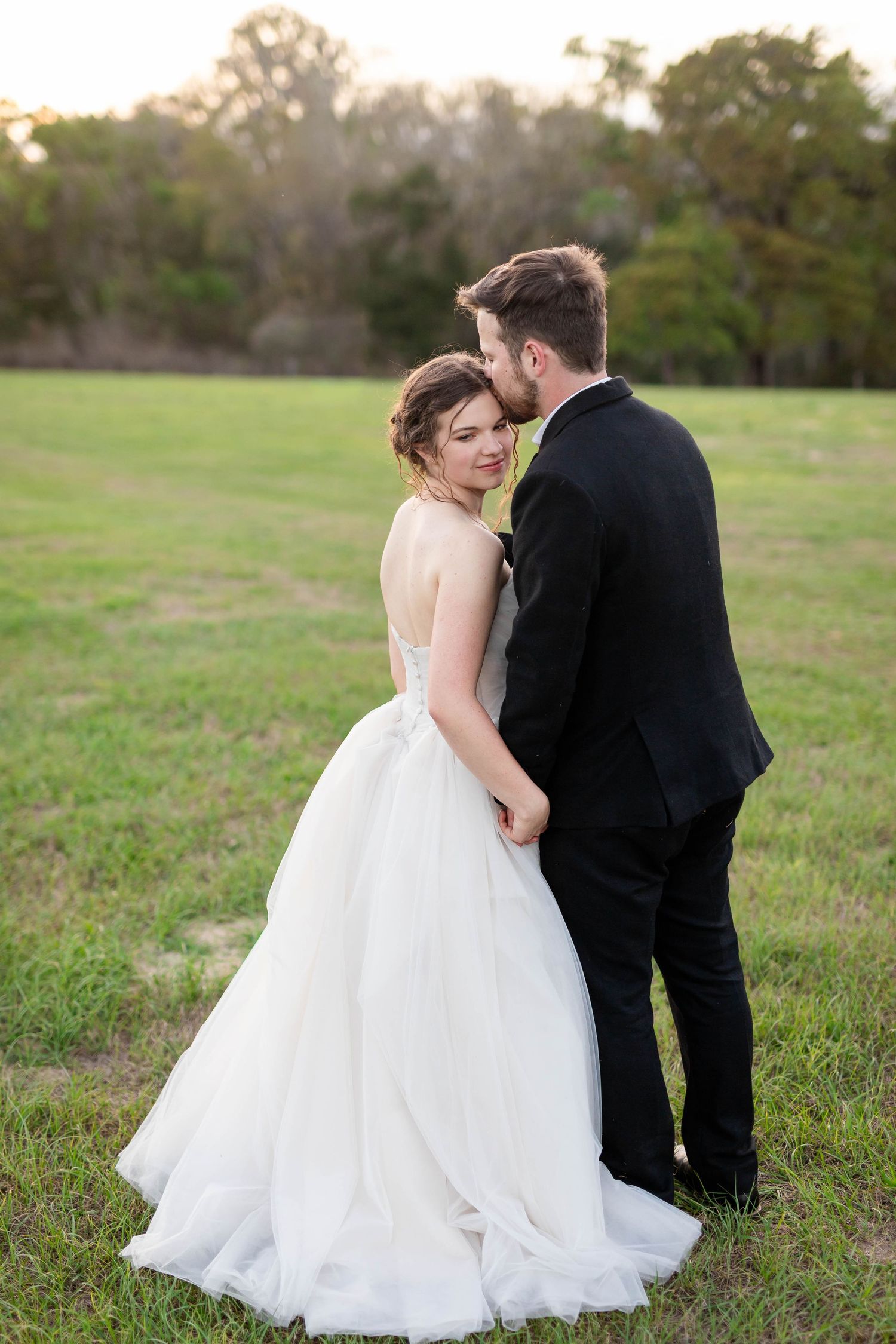 Bride in backless ballgown smiling over shoulder as groom kisses her cheek in a lush green field.