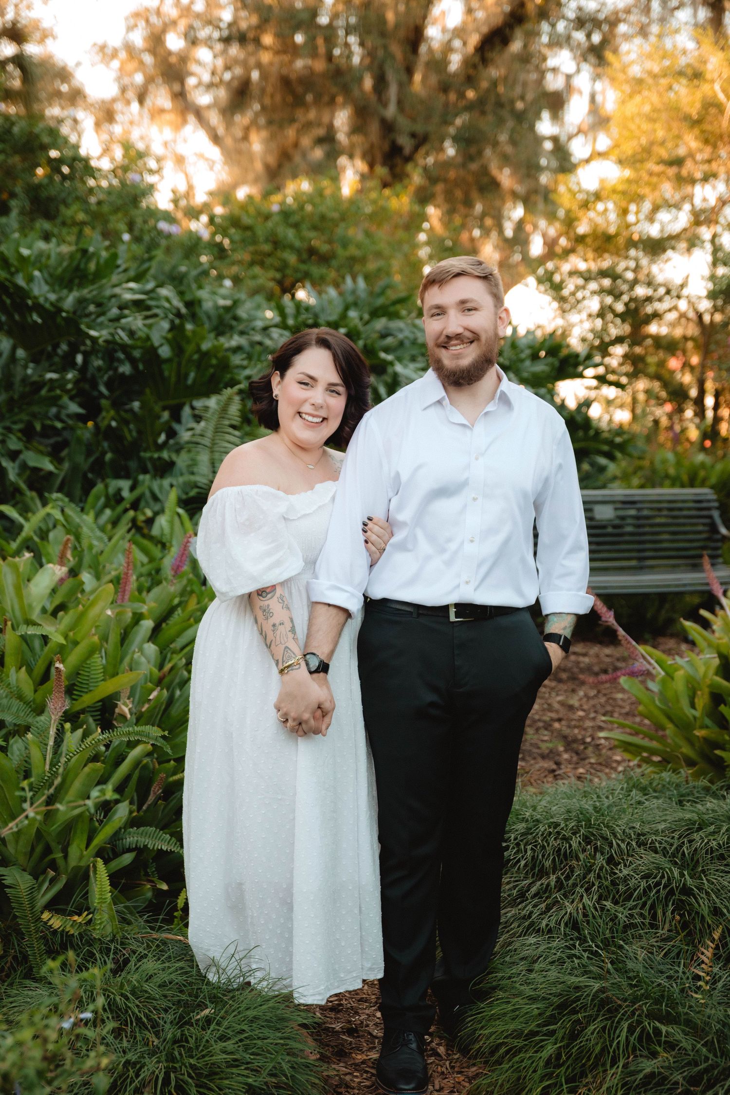 Smiling couple posing together in a lush garden at golden hour, woman wearing white off-shoulder dress at Bok Tower Gardens, Lake Wales.