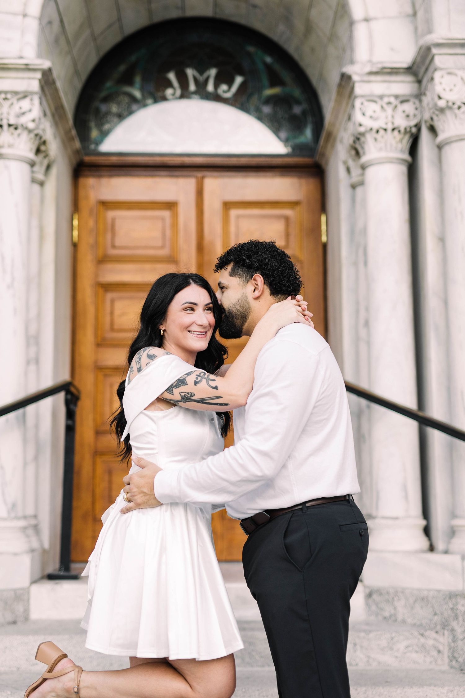 Couple embracing and smiling in front of ornate church doors with JMJ monogram above entrance in Tampa.