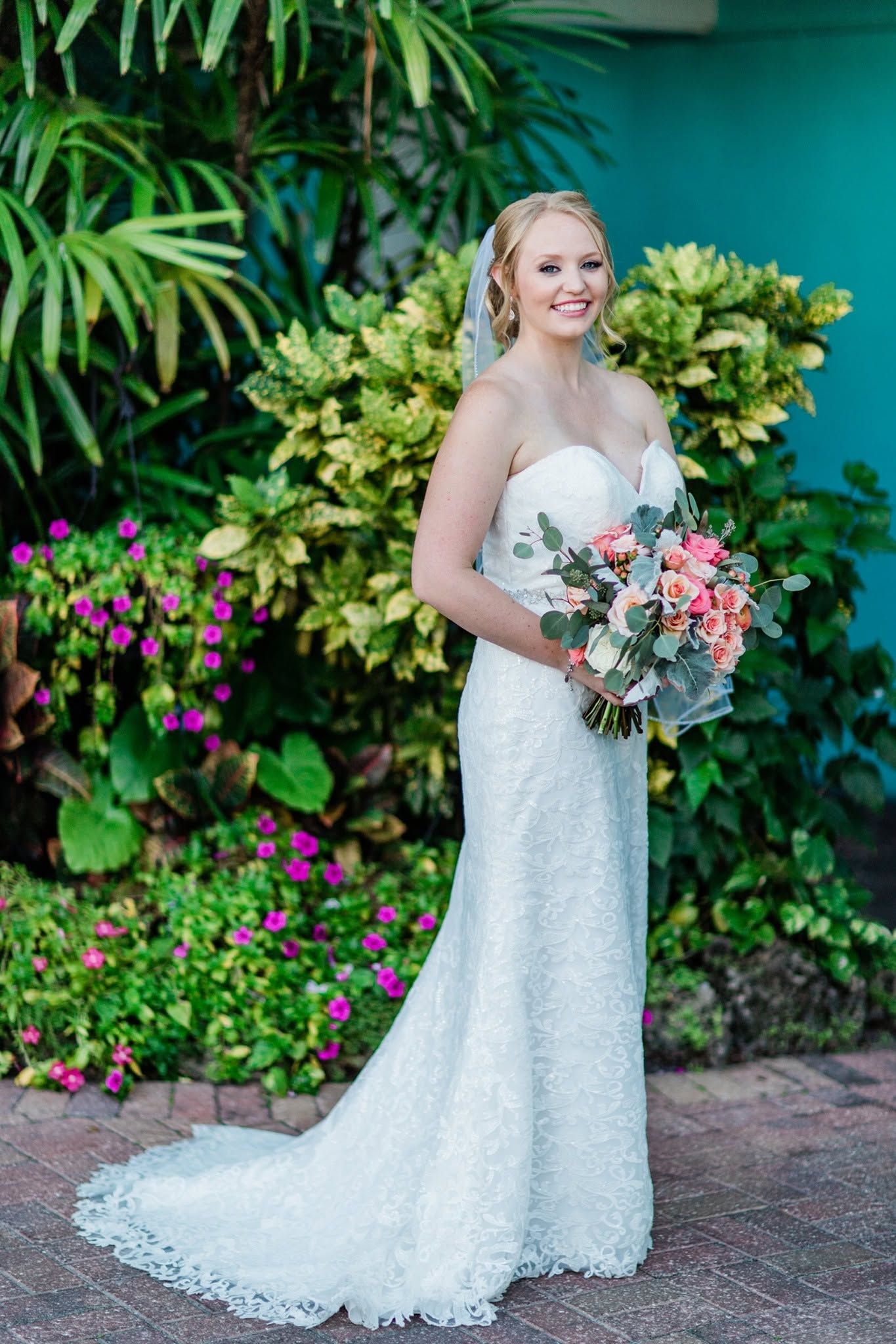Smiling bride in strapless lace gown holds pink bouquet before vibrant tropical garden backdrop.