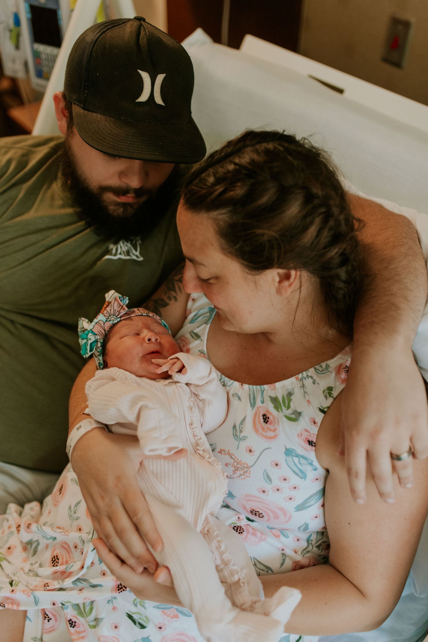 New parents in hospital room lovingly gazing at their newborn baby girl in floral outfit.
