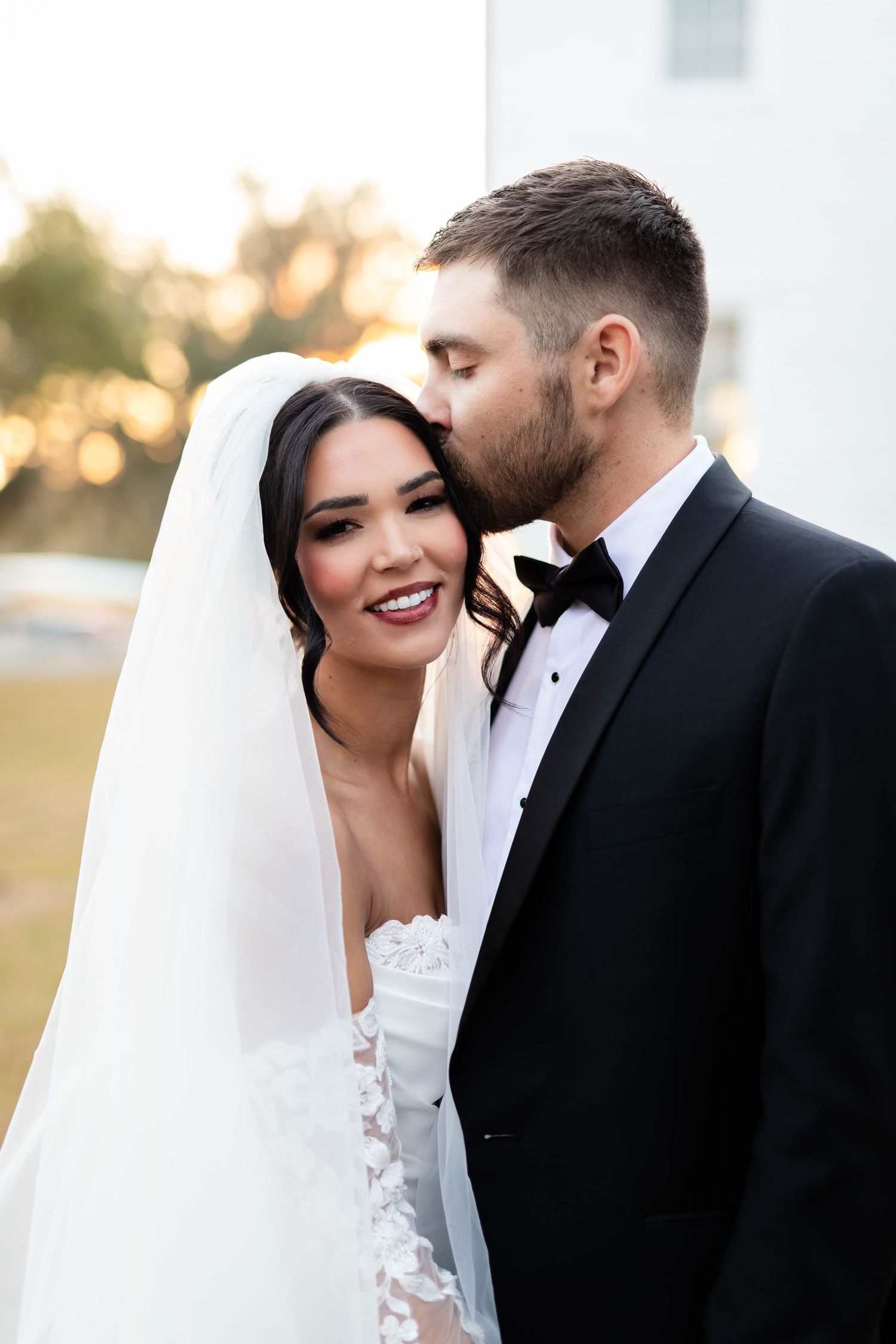 Groom in black tuxedo tenderly kisses bride in lace veil on the cheek during golden hour portraits in Tampa.