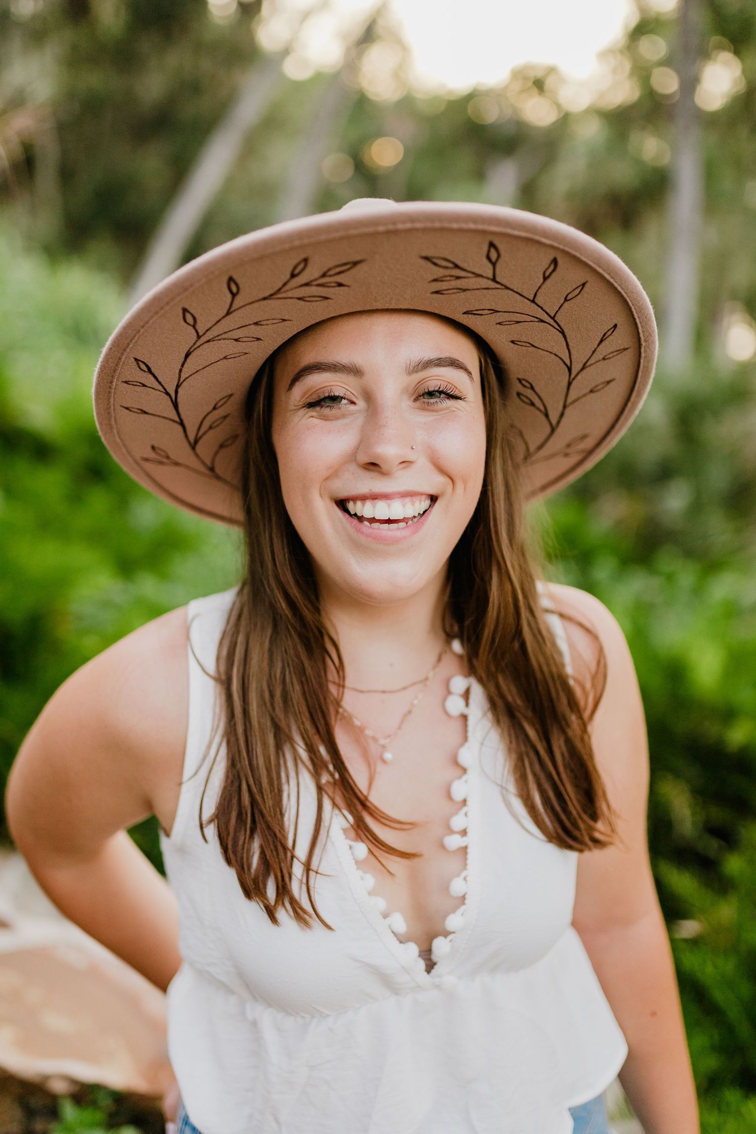 Smiling young woman wearing a tan wide-brim hat with floral design, white top, and layered necklaces outdoors.