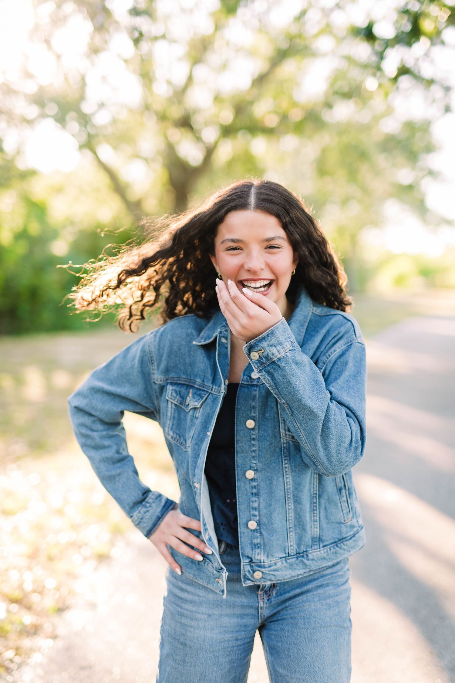 Young woman in denim jacket laughing outdoors with curly hair blowing in the wind on a sunny day.