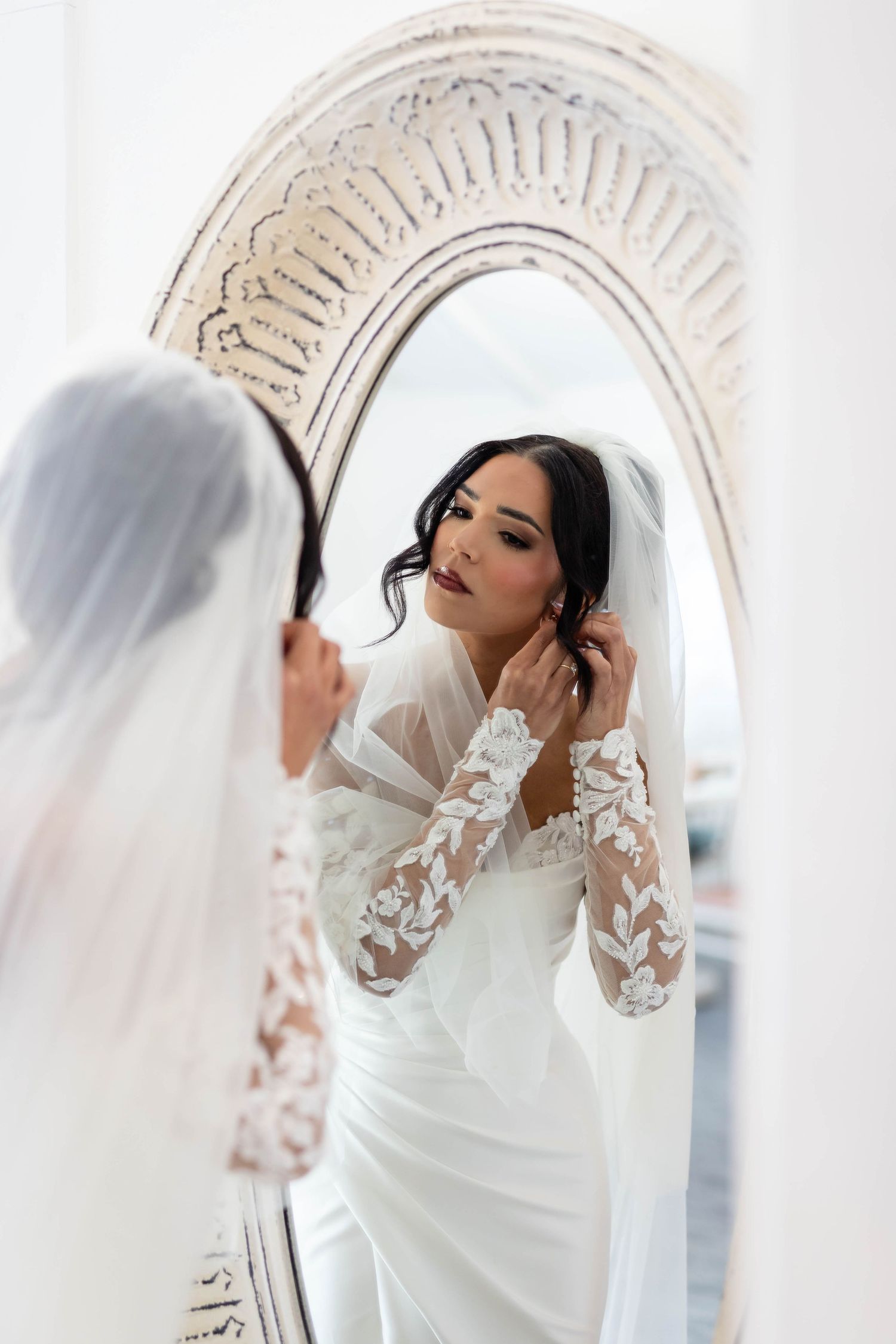 Bride in lace-sleeved white gown adjusts earring while gazing at her reflection in an ornate arched mirror.