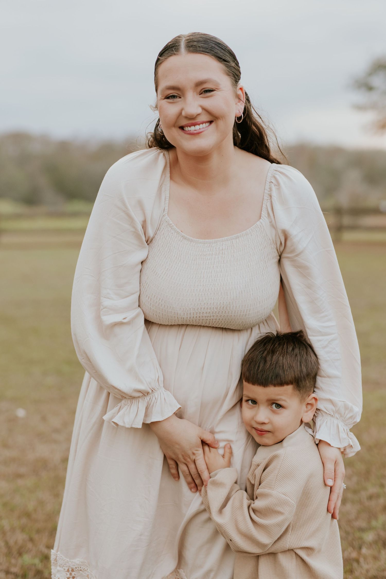 Smiling mother in cream dress standing outdoors with young son hugging her waist.