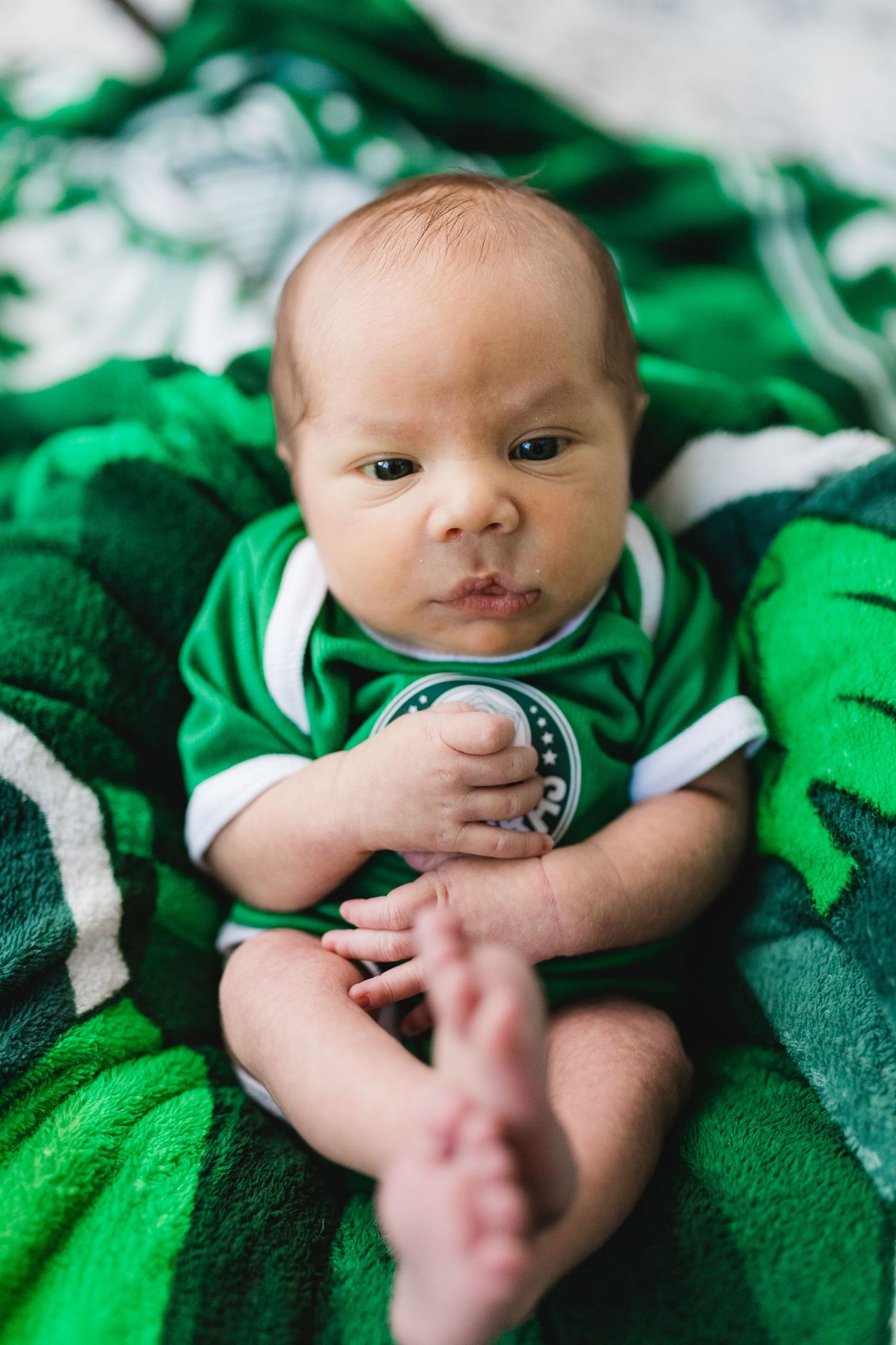 Newborn baby in green soccer jersey lying on green blanket looking at camera.