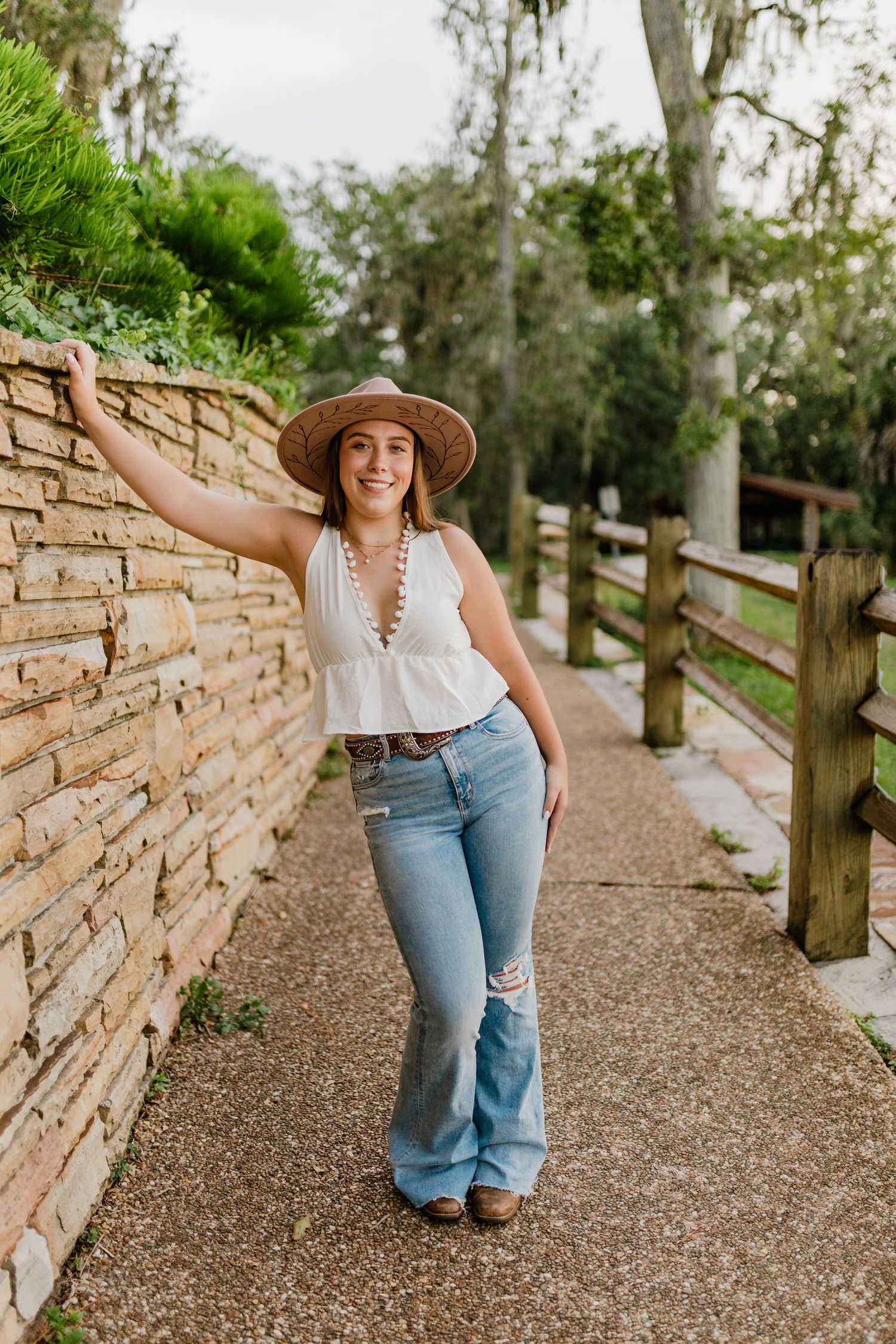 Young woman in wide-brim hat, white top, and flare jeans poses along stone wall on rustic pathway.
