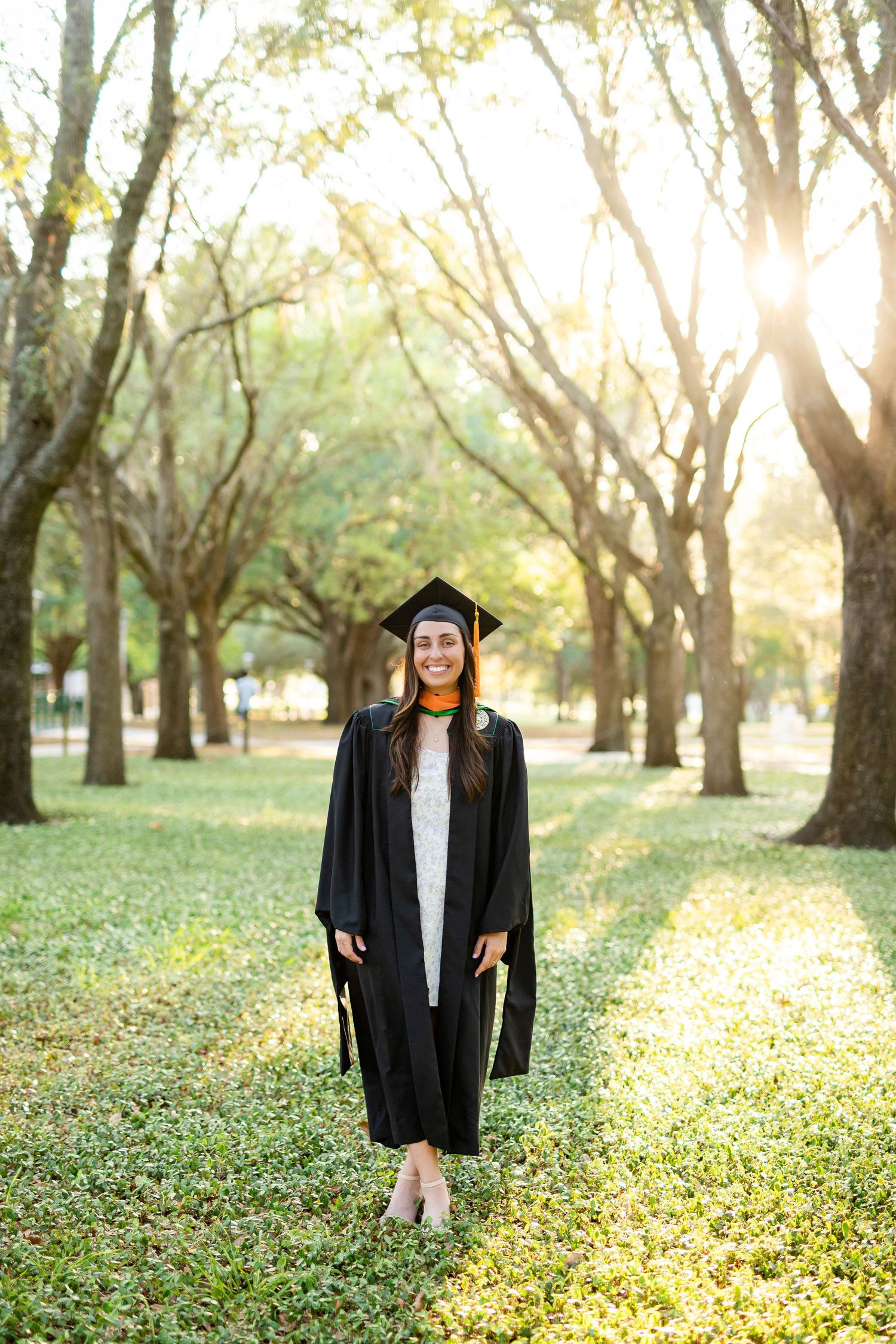 Smiling graduate in cap and gown stands in sunlit tree-lined park path at the Unviersity of South Florida.