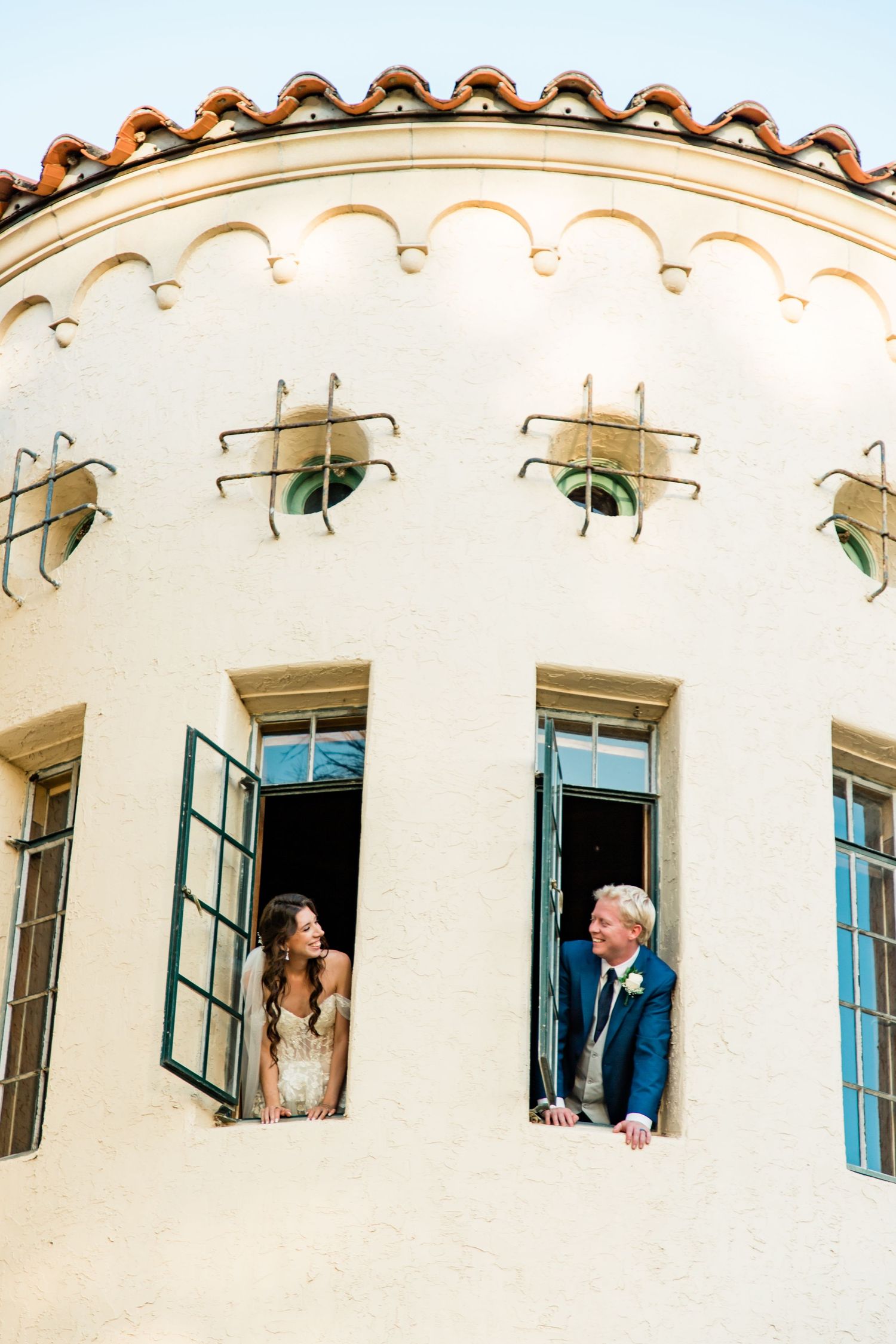 Bride and groom lean out of separate windows of a Spanish-style tower building, smiling at each other at Powel Crosley Estate, Sarasota.
