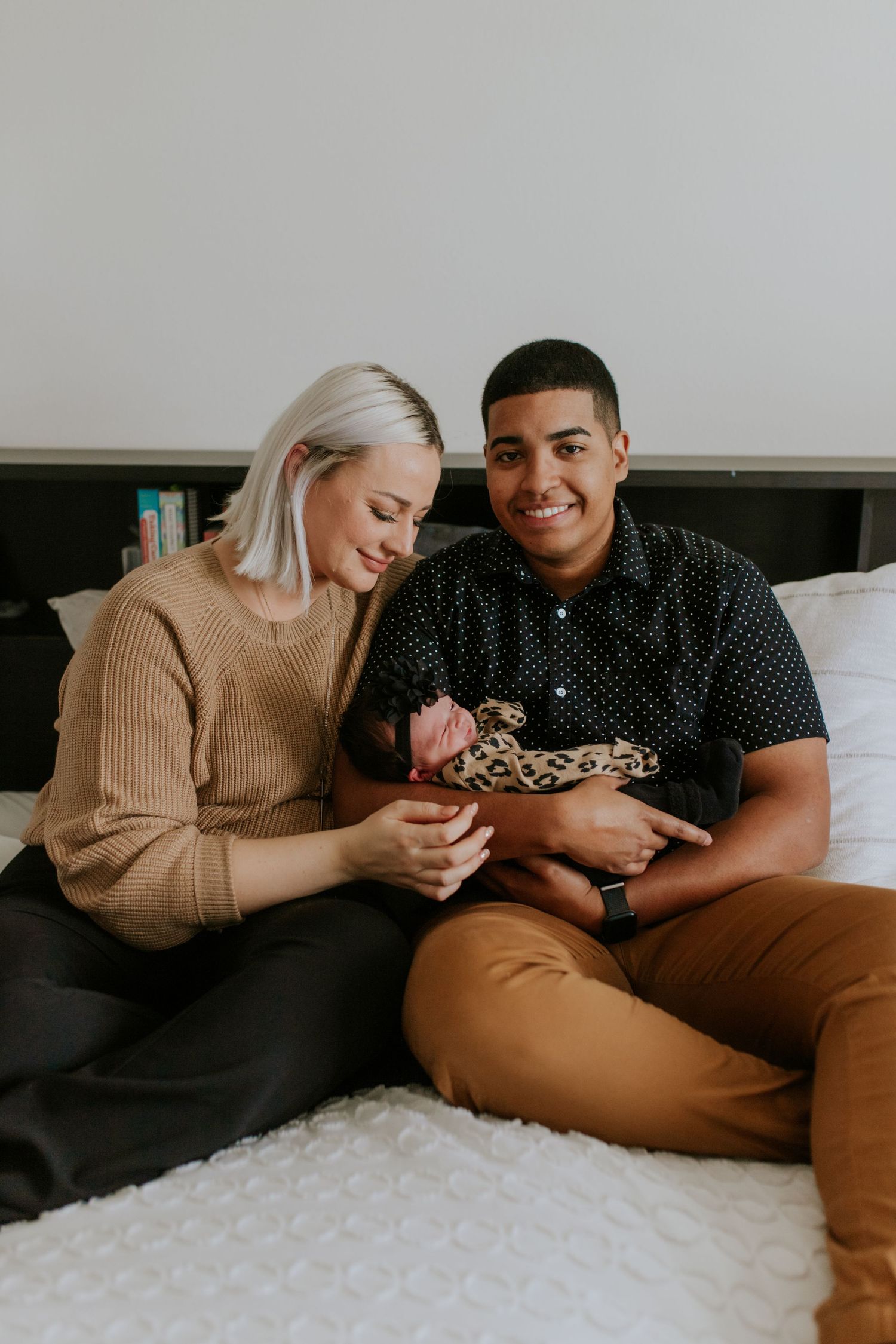 Smiling young couple sitting on bed together holding their newborn baby wrapped in leopard print.