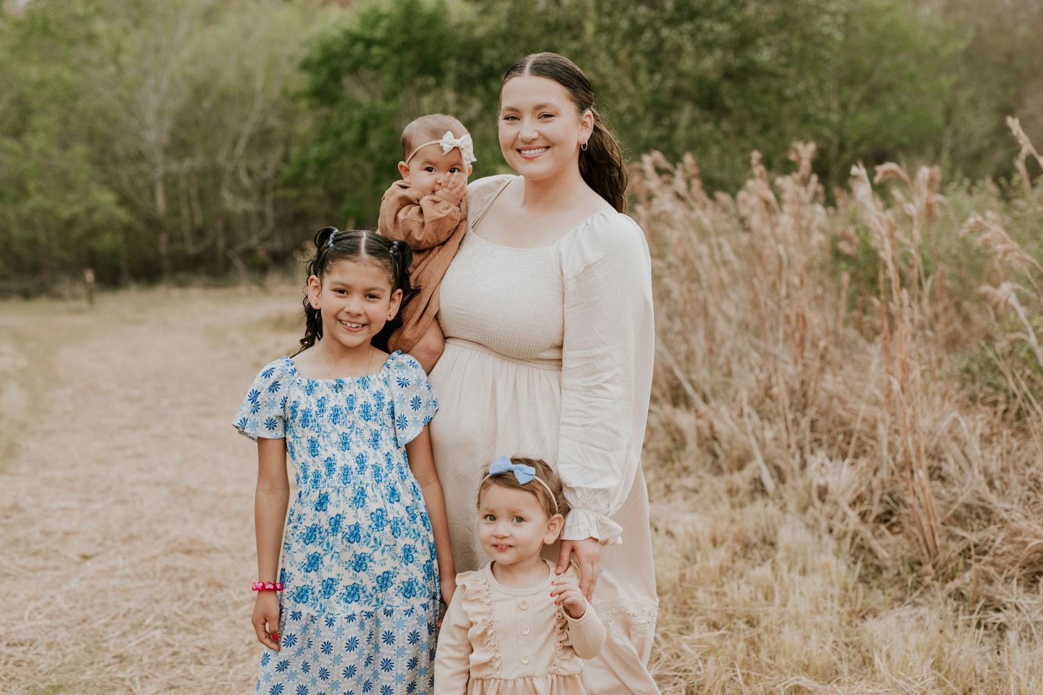 Smiling mother in white dress poses with three young daughters on a dirt path.