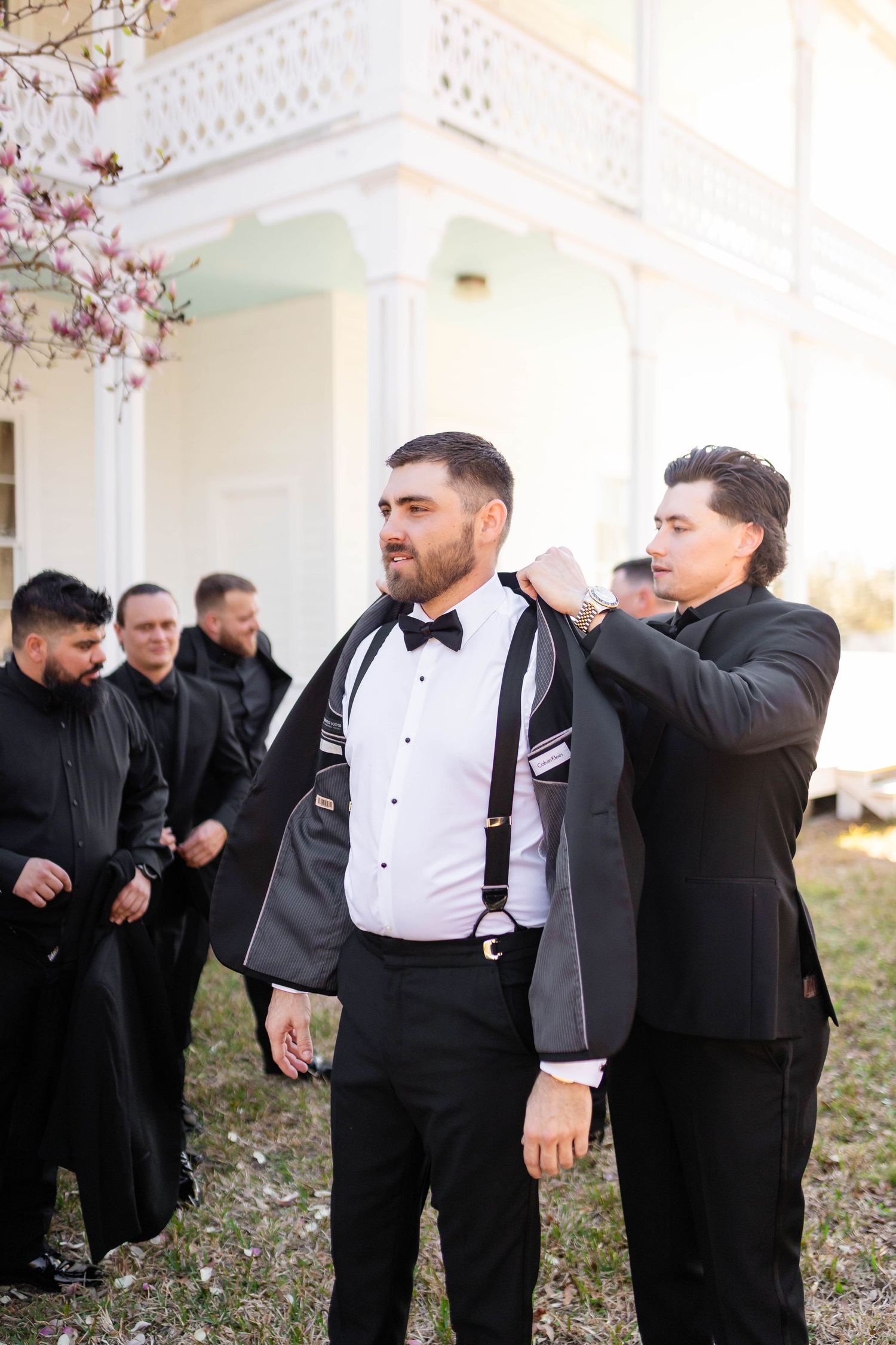 Groomsmen in black suits help groom with his jacket outside a white historic venue before the wedding in Tampa.