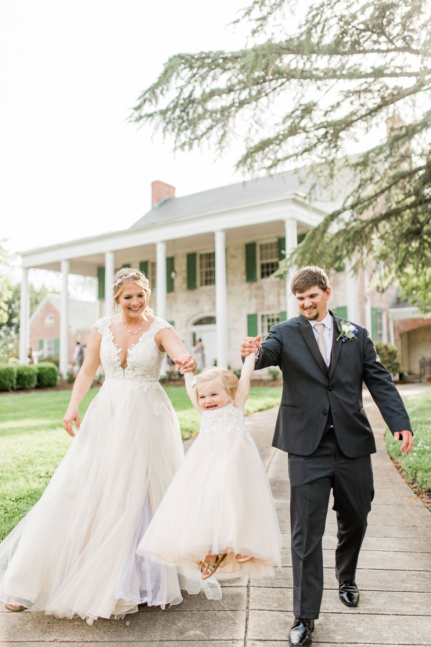 Smiling bride and groom swing young flower girl between them on garden pathway outside estate.