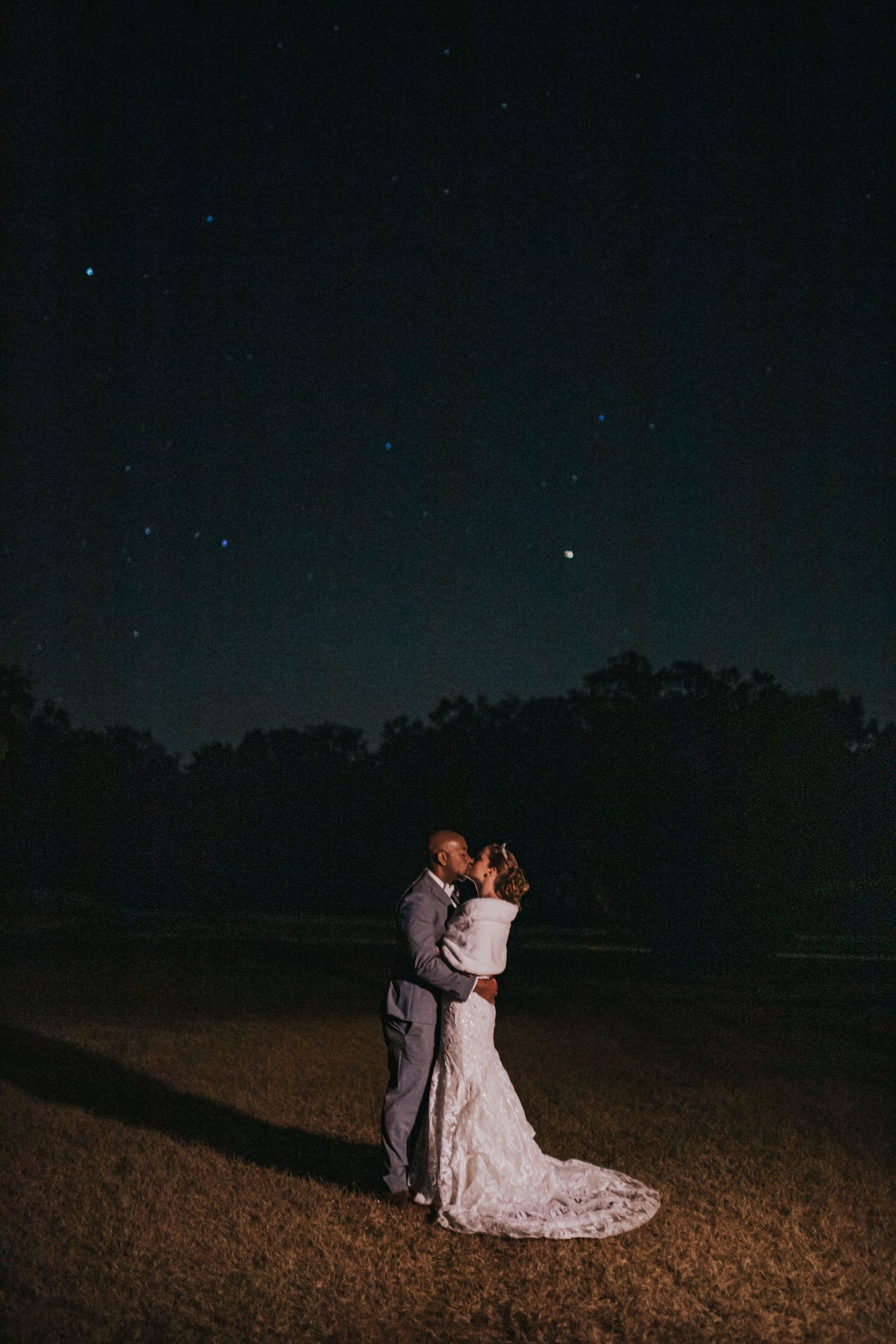Bride and groom share a romantic kiss under a stunning starry night sky at their outdoor wedding at Legacy Lane.