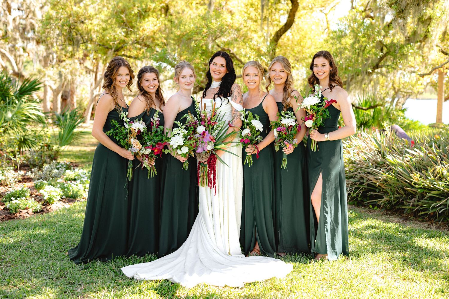Bride poses with six bridesmaids in emerald green gowns holding tropical bouquets in a lush garden.