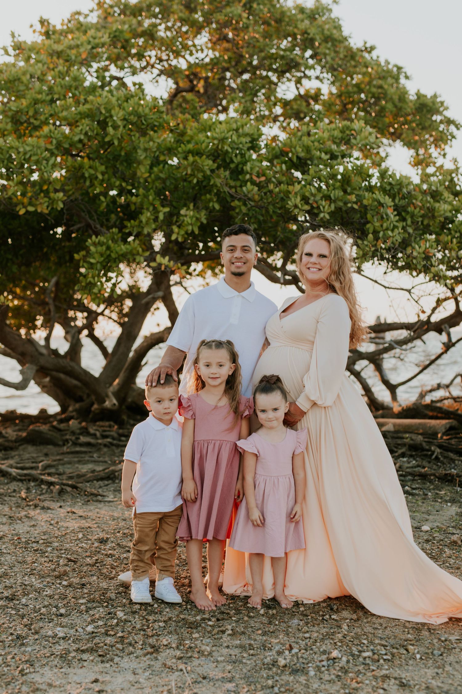 Pregnant woman with family including three children posing under a tree at beach.